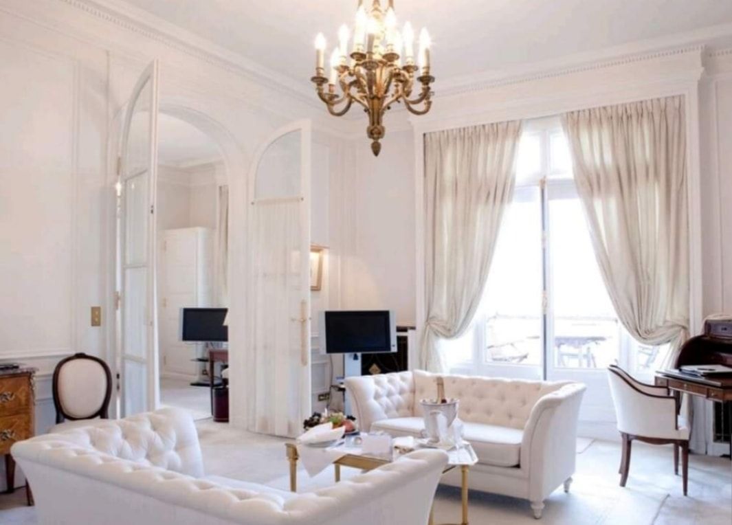 Elegant white living room with tufted sofas, chandelier, arched doorway, and large window with drapes