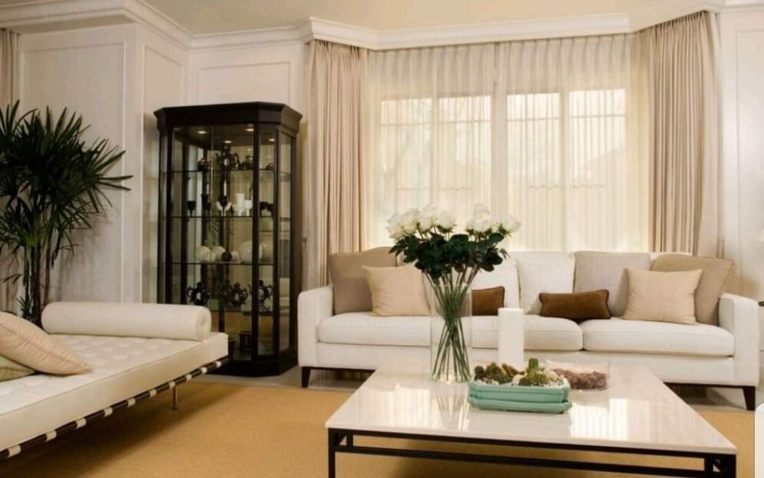 Bright living room with white sofas, glass cabinet, coffee table, and large sheer-curtained windows