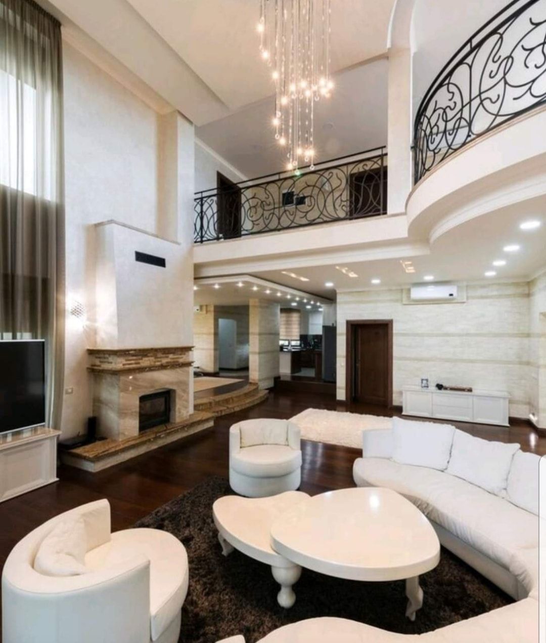 Spacious modern living room with white curved sofas, fireplace, balcony, and chandelier lighting