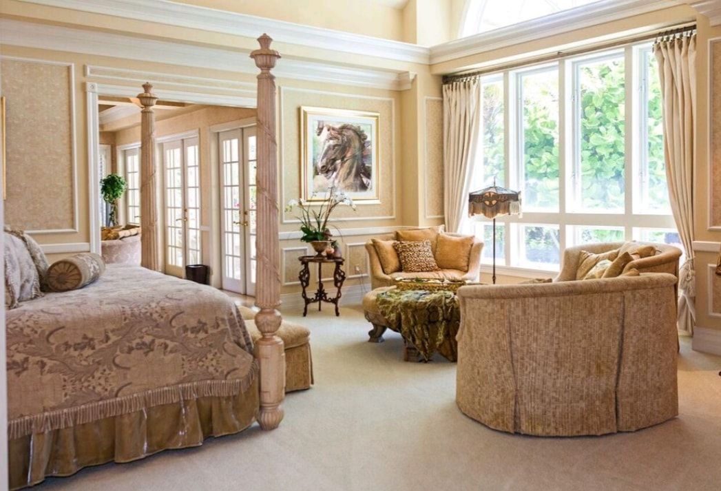 Elegant cream-toned bedroom with a canopy bed, seating area, large windows, and ornate decor