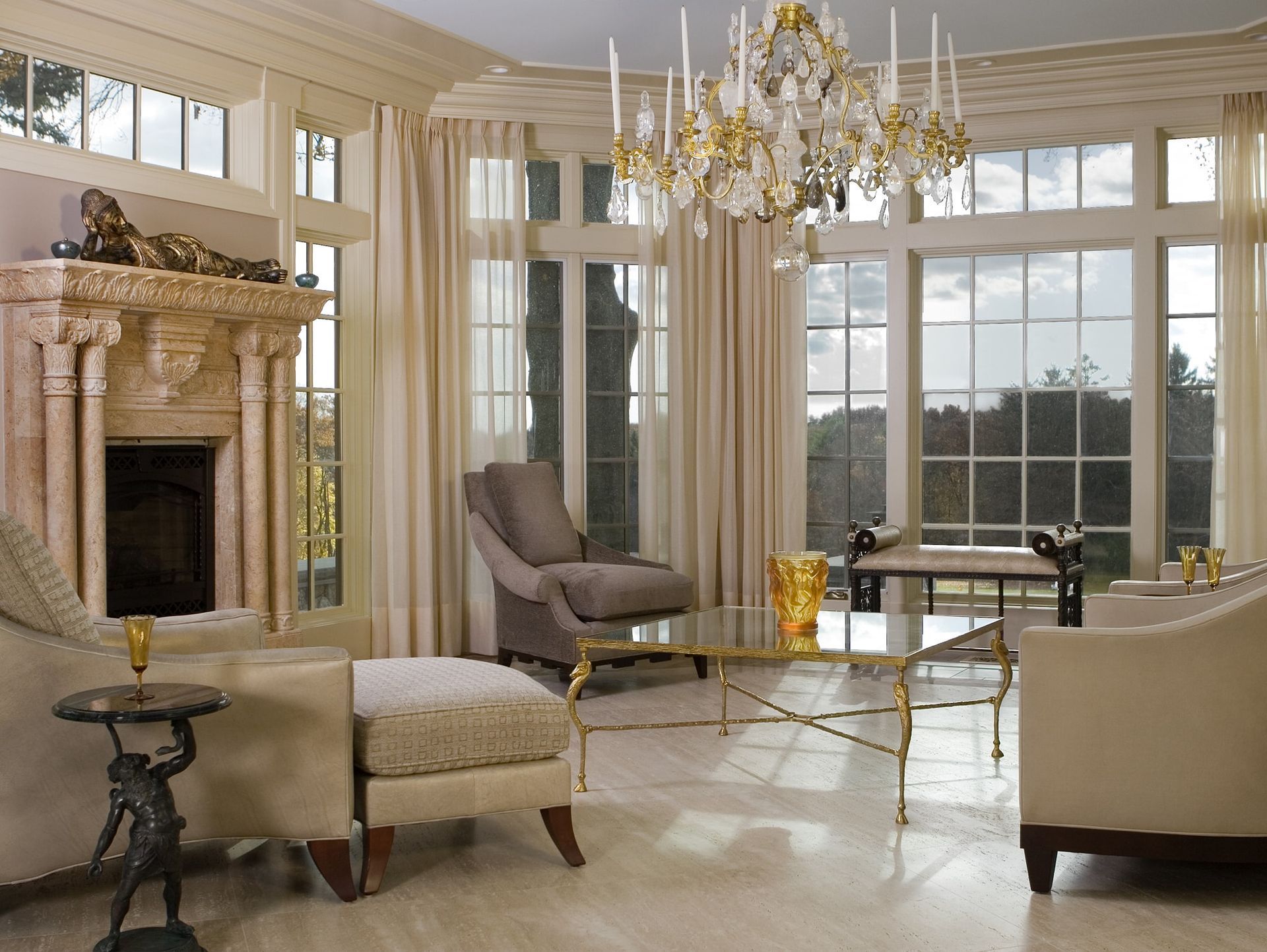 Elegant living room with fireplace, chandelier, cream chairs, and large windows overlooking greenery