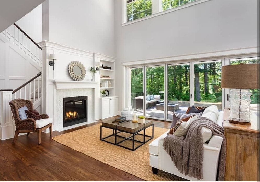Bright living room with white fireplace, hardwood floors, gray sofa, and large windows overlooking greenery