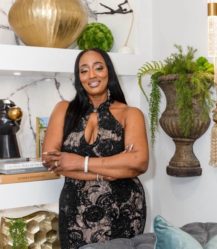 Woman in black lace dress standing in a stylish living room beside shelves and a large urn planter.