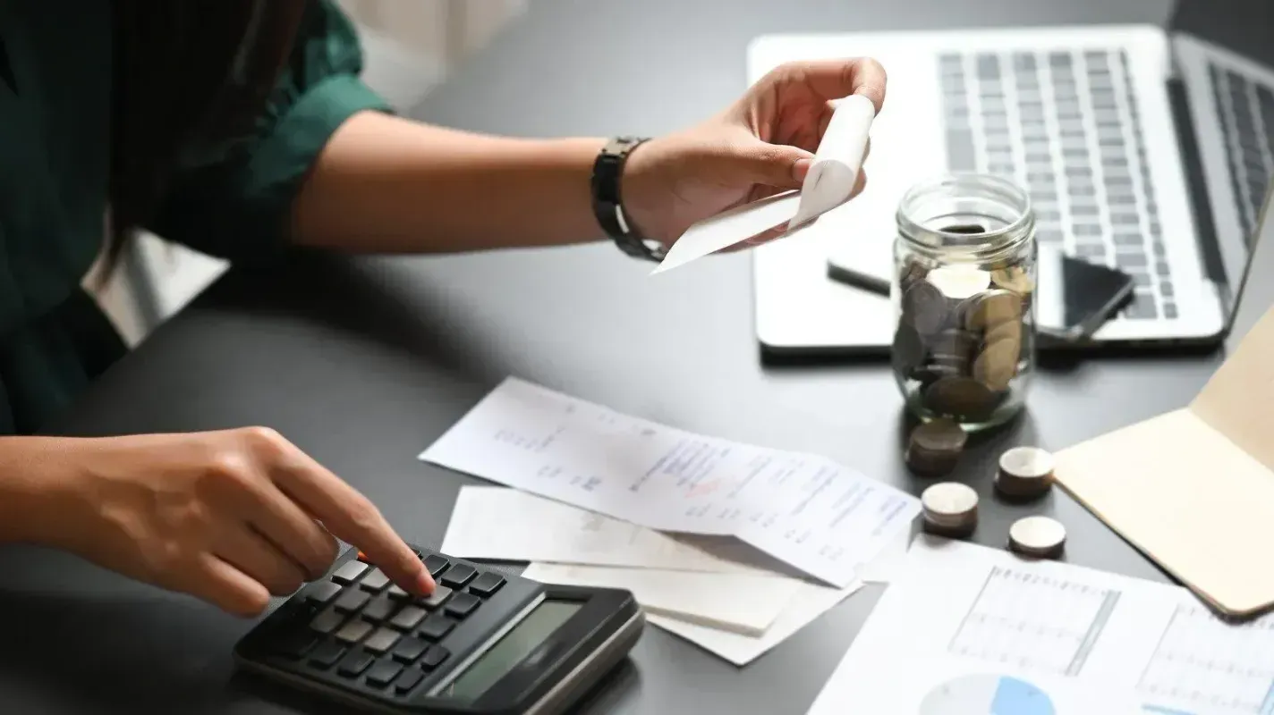 Hands using a calculator and paper receipts beside a laptop and jar of coins on a desk