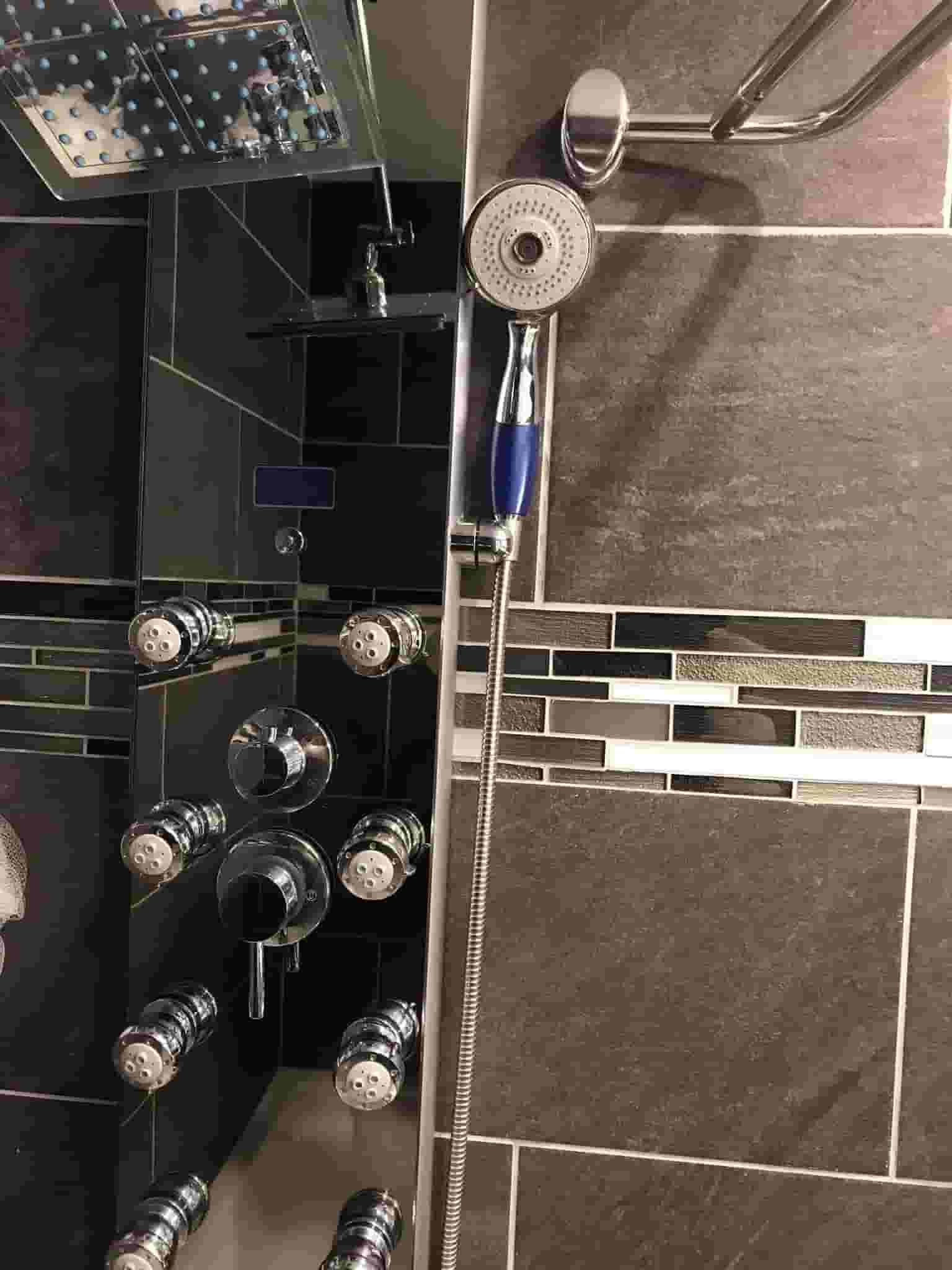 Glass shower with chrome showerhead, blue handle, and tiled walls; several bottles on a shelf.