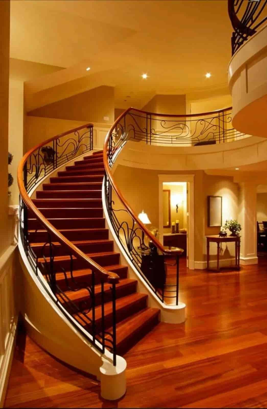 Grand curved staircase with red carpet, dark railings, and warm lighting in an elegant foyer
