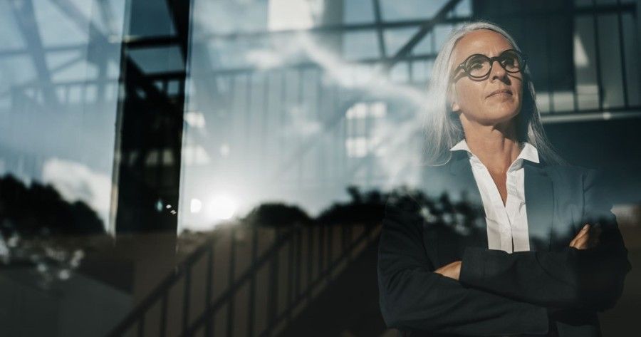 A woman in a suit and glasses is standing with her arms crossed in front of a building.