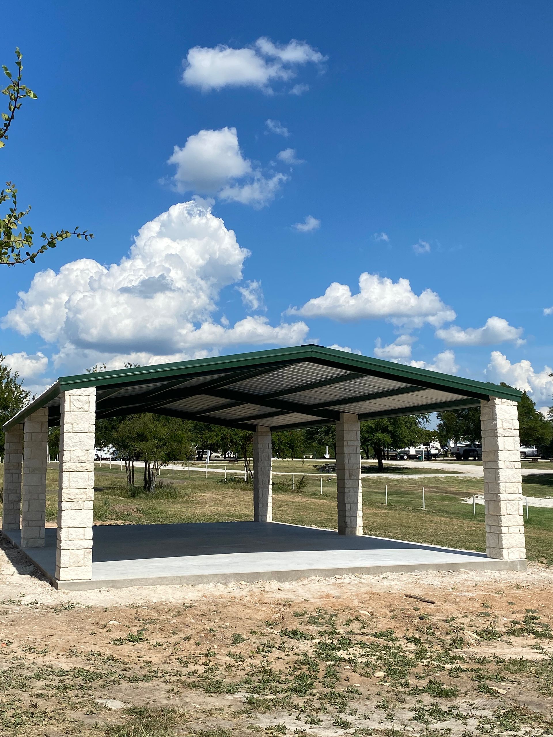 Open-sided stone pavilion with a green roof on a sunny grassy hillside under a blue sky