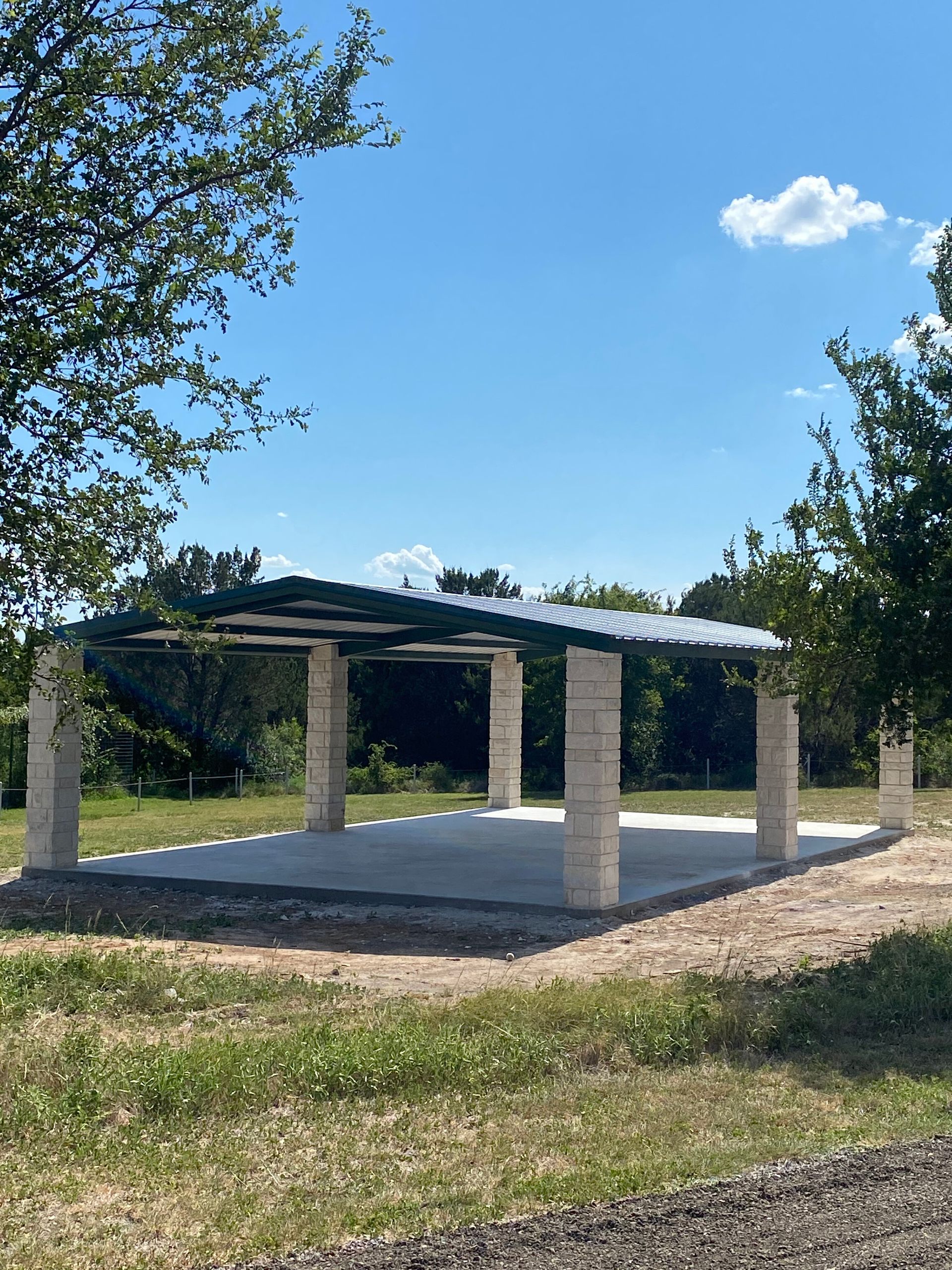 Outdoor pavilion with a white roof and stone pillars beside a grassy path under a blue sky
