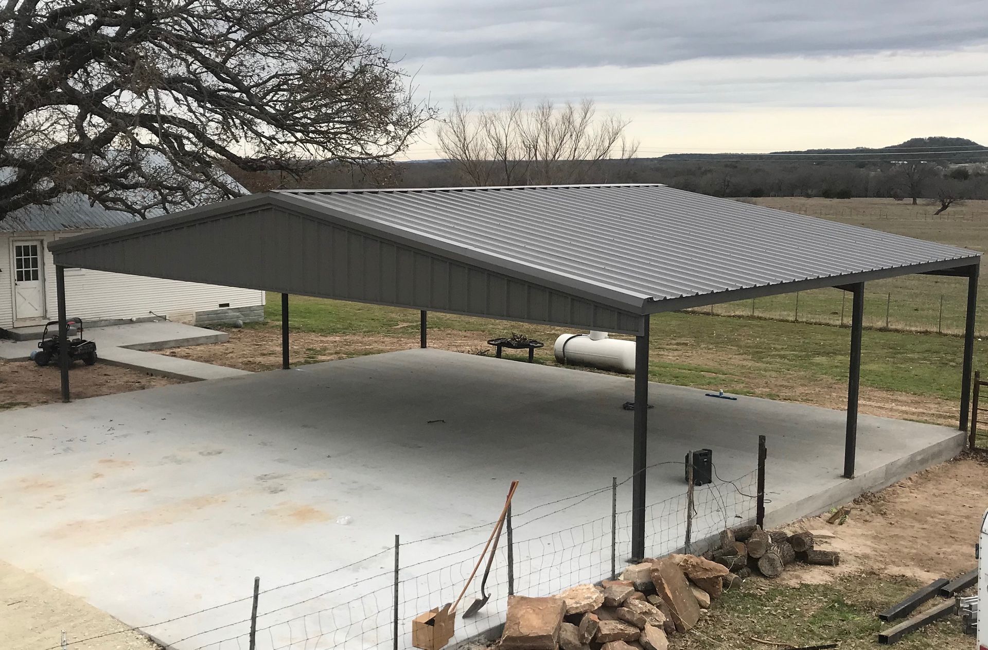 Steel-roofed open carport on a concrete pad with chain-link fencing in a rural yard.