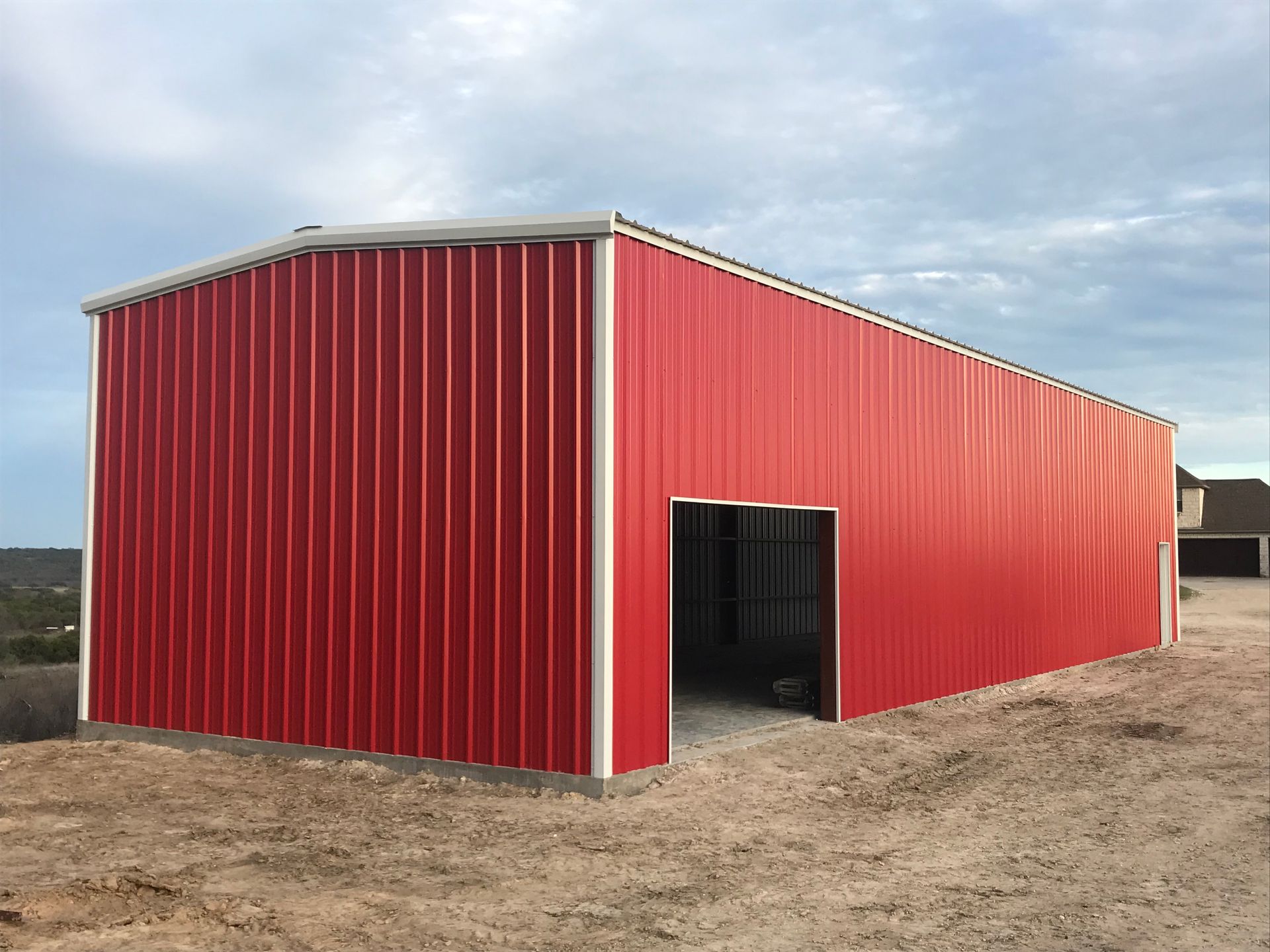 Red metal barn with an open doorway on a dirt lot under a cloudy sky
