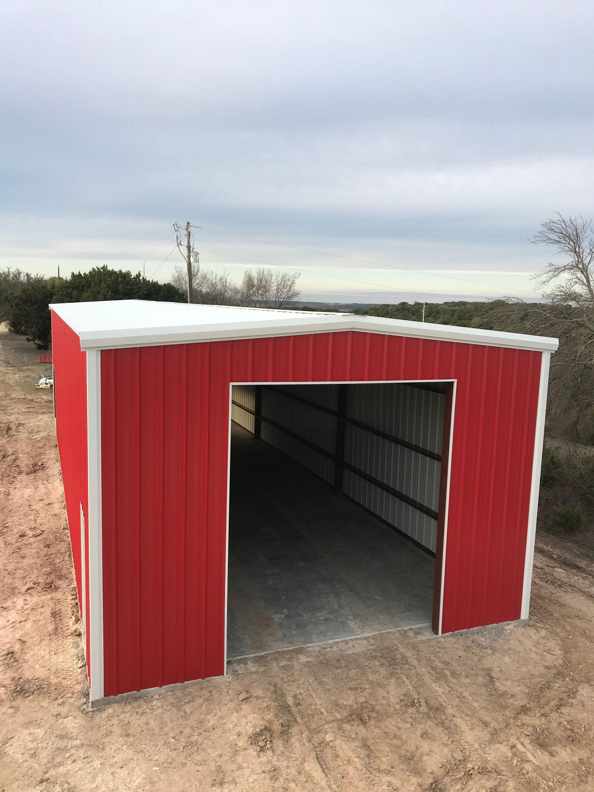 Small red metal shed with an open front in a dirt field under a cloudy sky