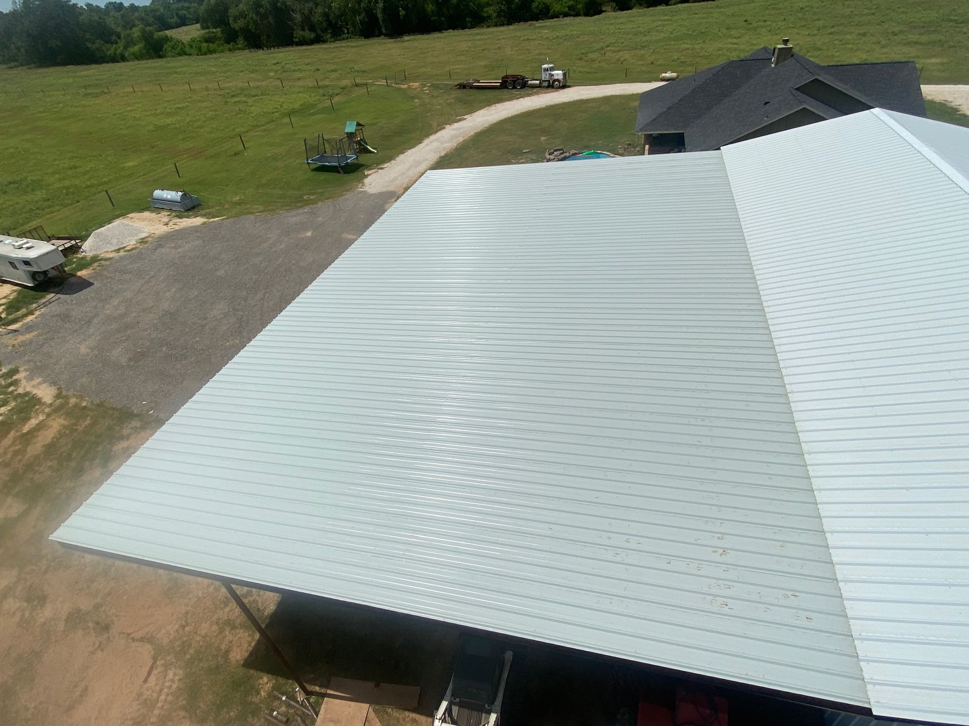 Aerial view of a large white metal-roofed building beside a gravel driveway and grassy field.