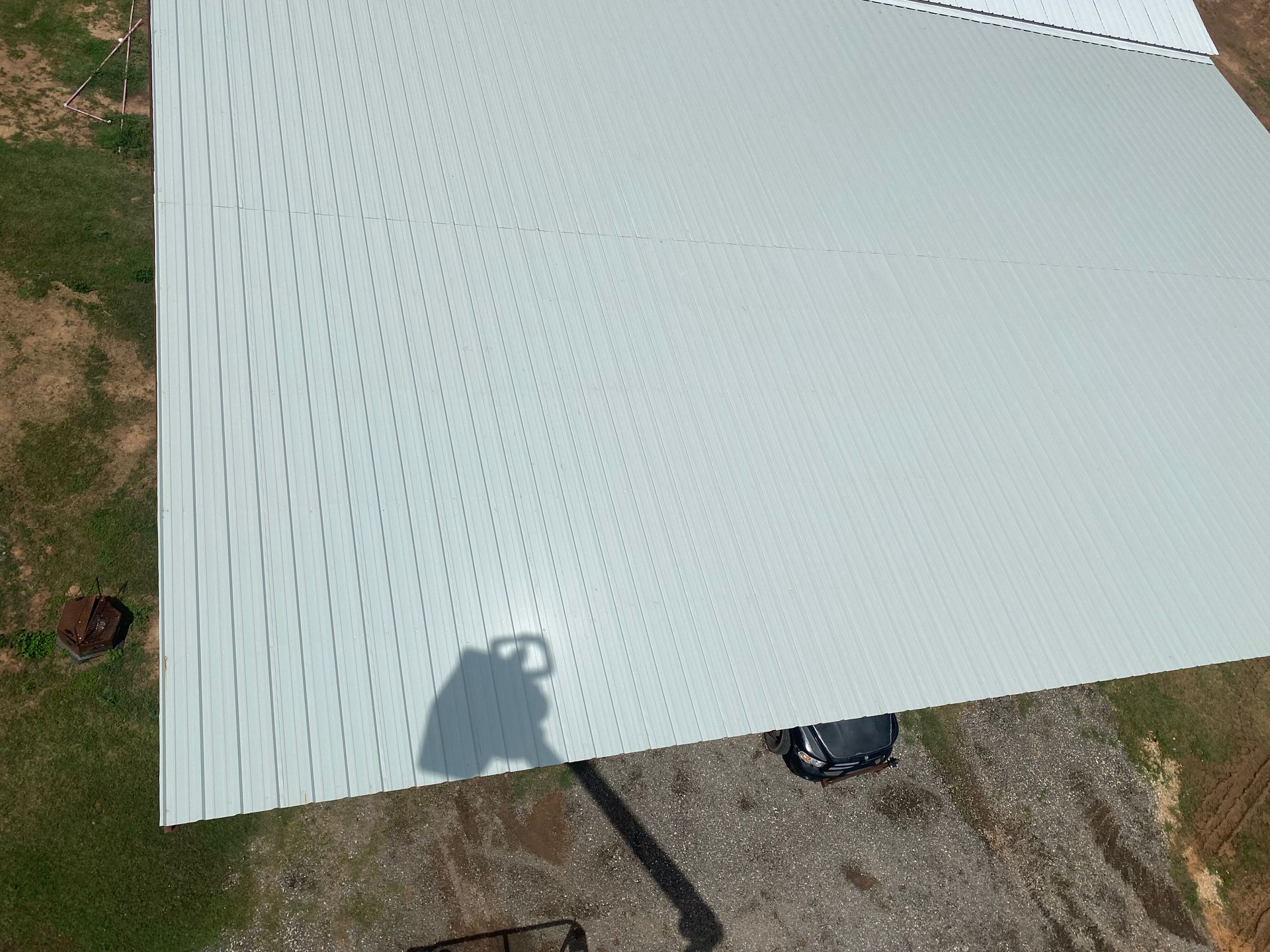 Aerial view of a light gray corrugated metal roof beside patchy grass and a small dark vehicle.