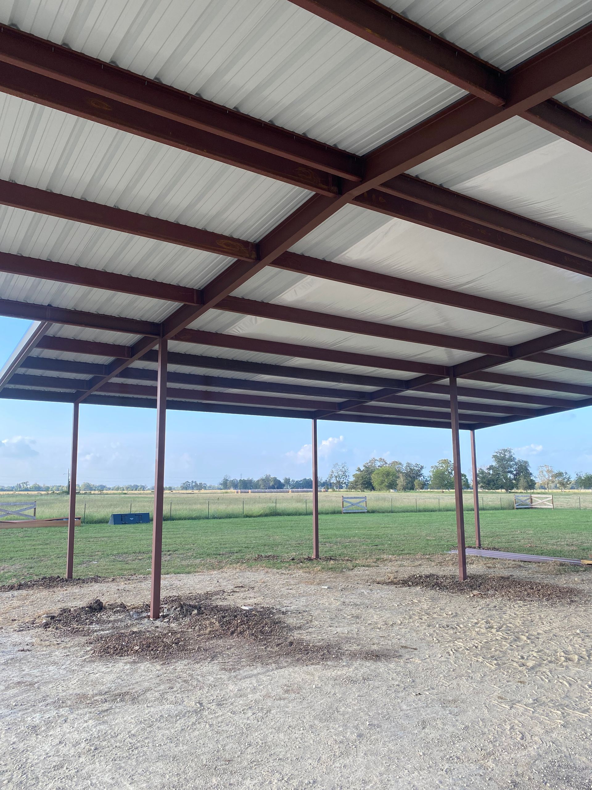 Open-sided metal shed with red beams over a gravel lot and grassy field beyond