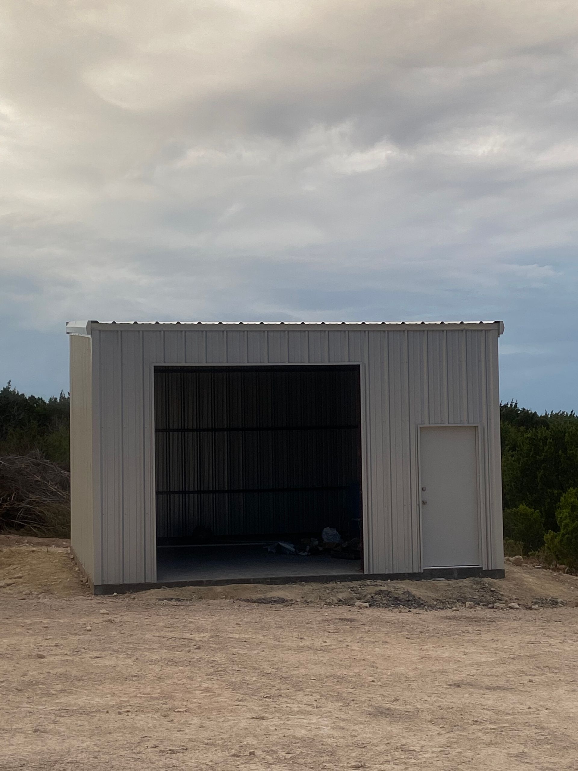 Small white metal shed with an open front and closed side door on a dirt lot under a cloudy sky