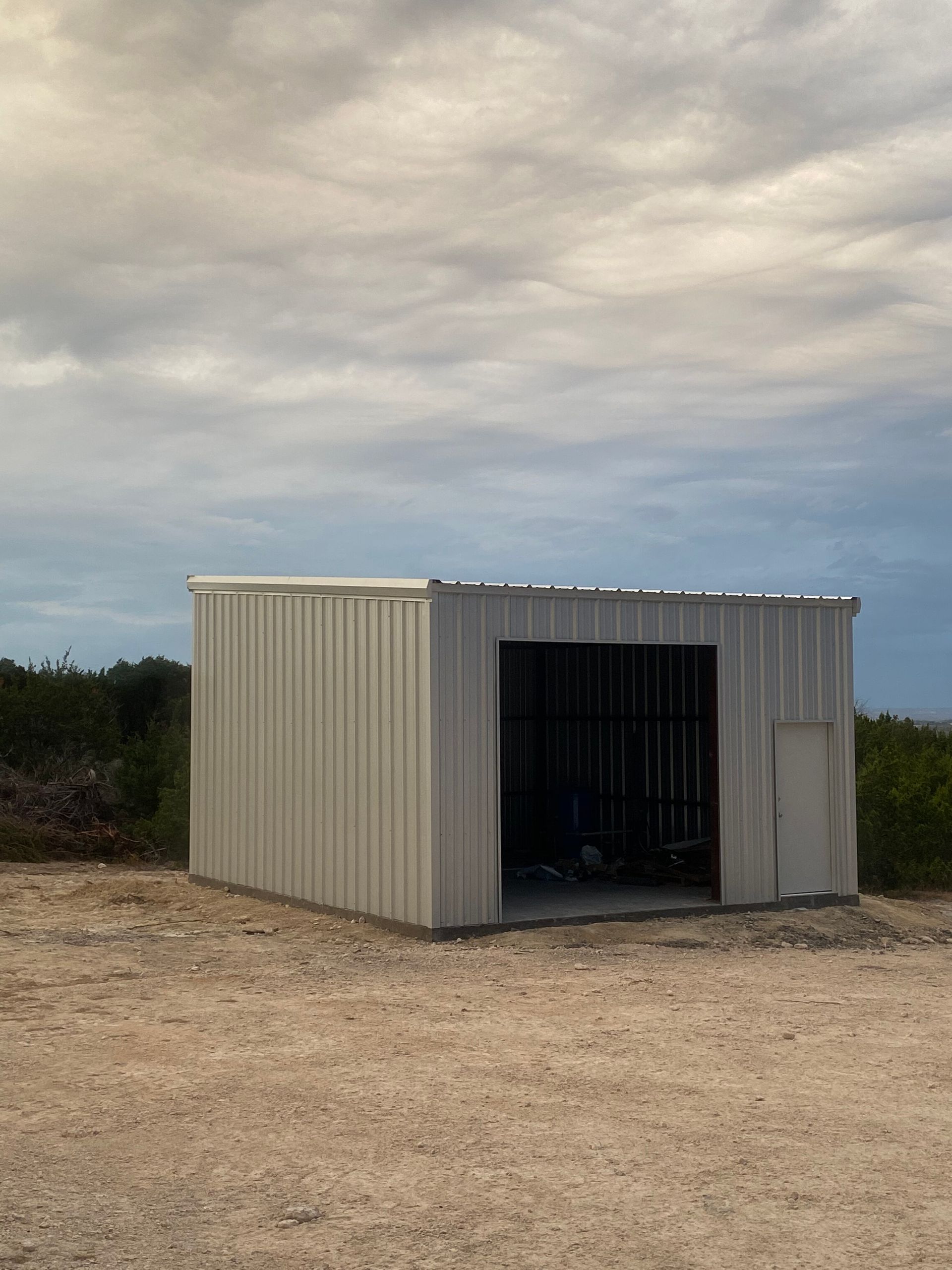 Small beige metal shed with open doorway on a dirt lot under a cloudy sky
