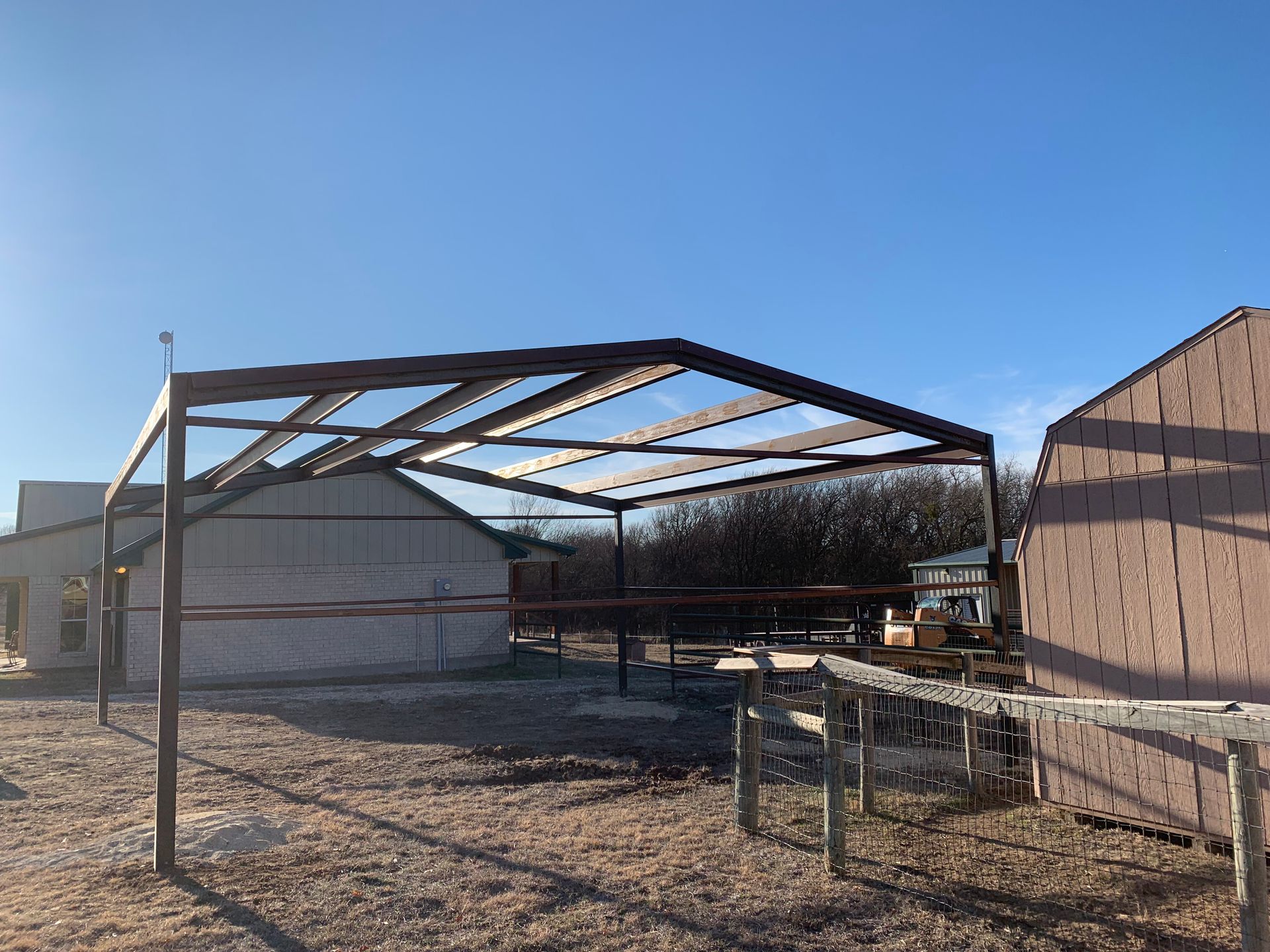 Partially built metal shed frame in a gravel yard beside tan buildings under a blue sky