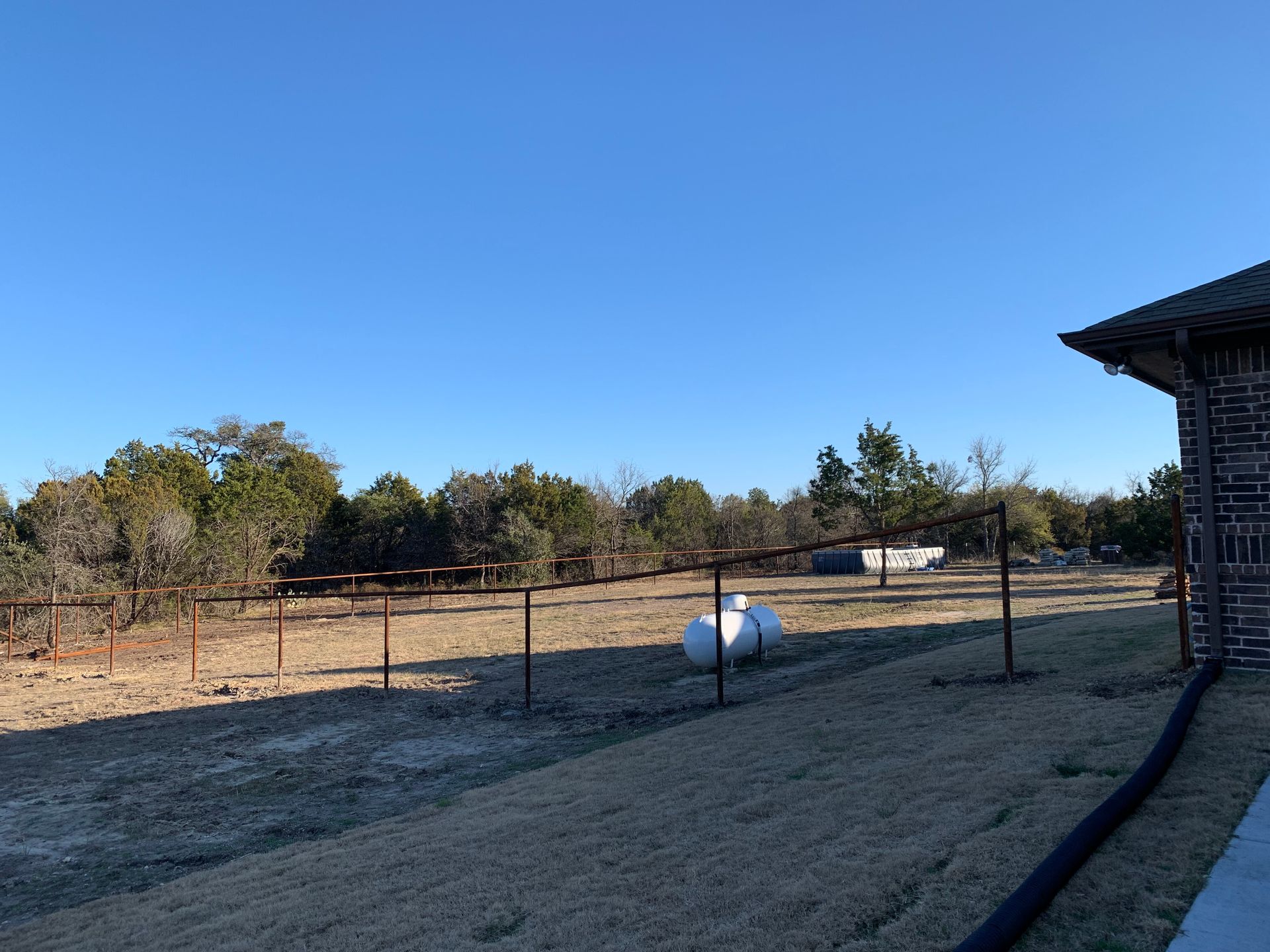 Rural fenced yard with a white propane tank, dry grass, trees, and a clear blue sky