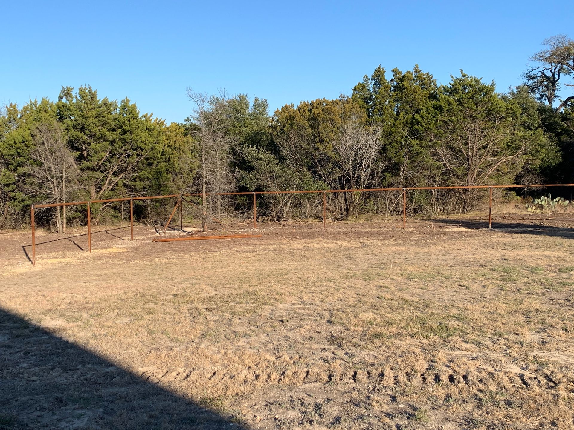 Dry fenced dirt clearing with tire tracks, backed by trees under a clear blue sky