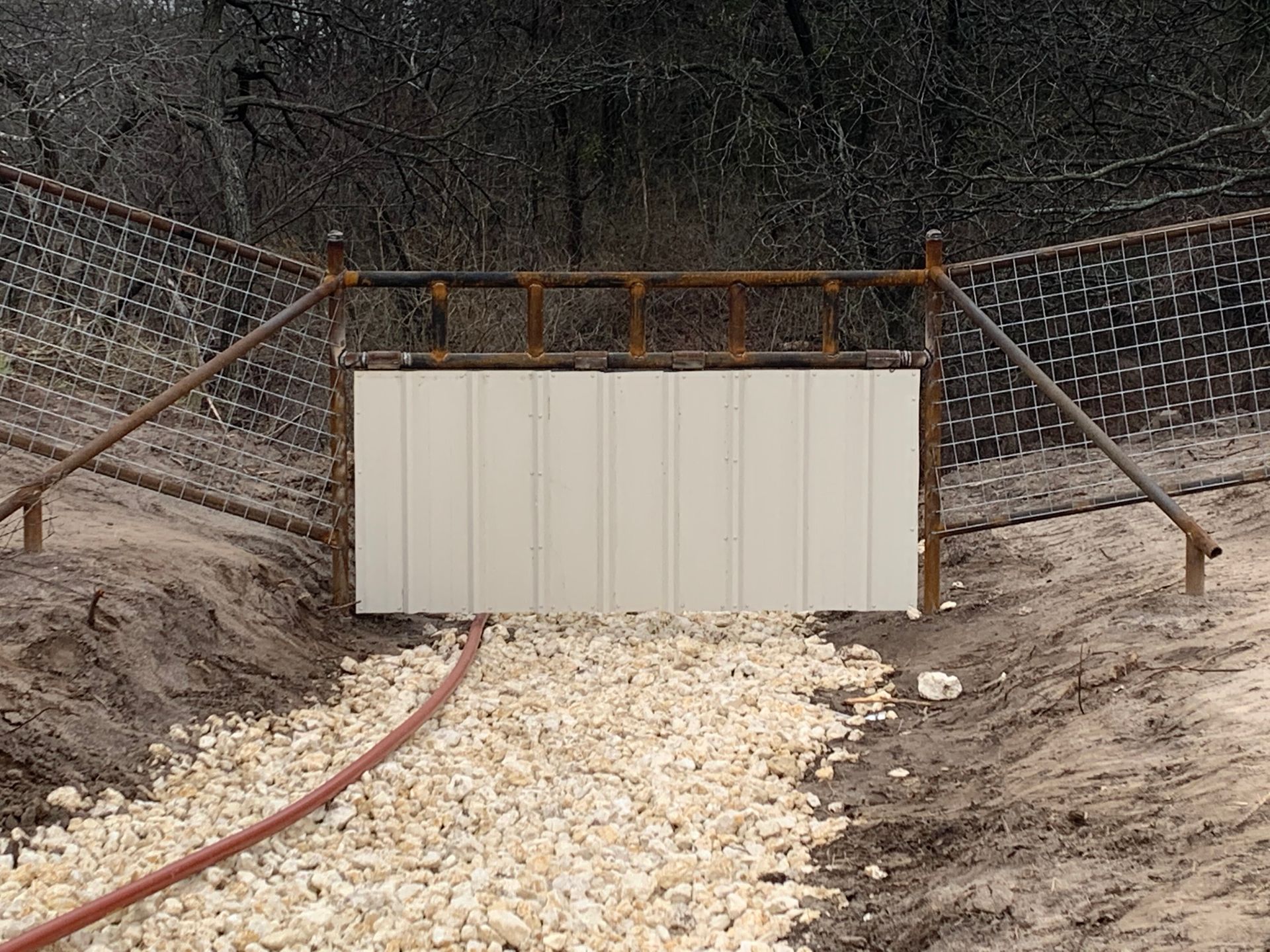 Closed beige metal gate across a gravel road in a wooded area