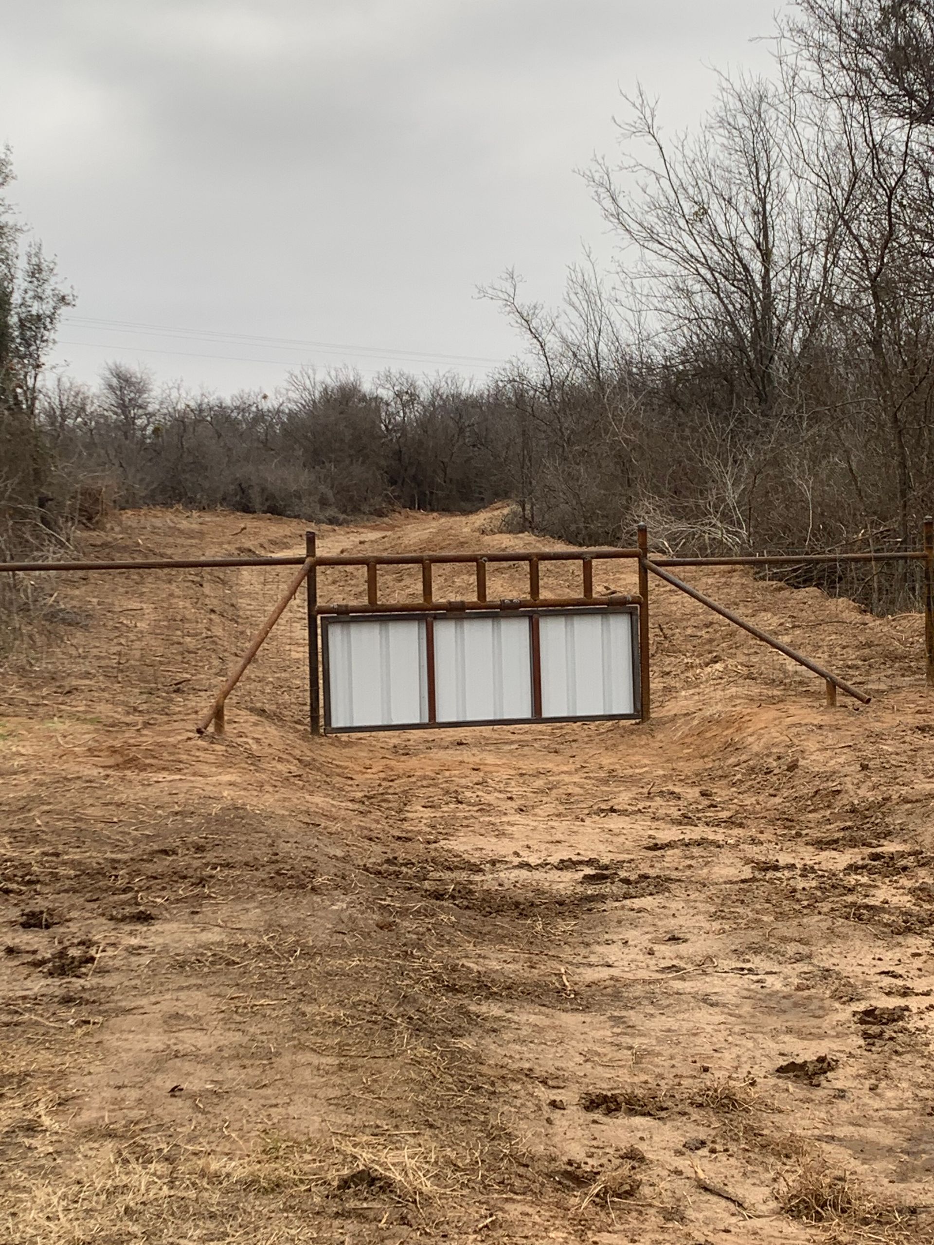 Blocked dirt road with a wooden-and-metal barrier across it in a dry, scrubby landscape