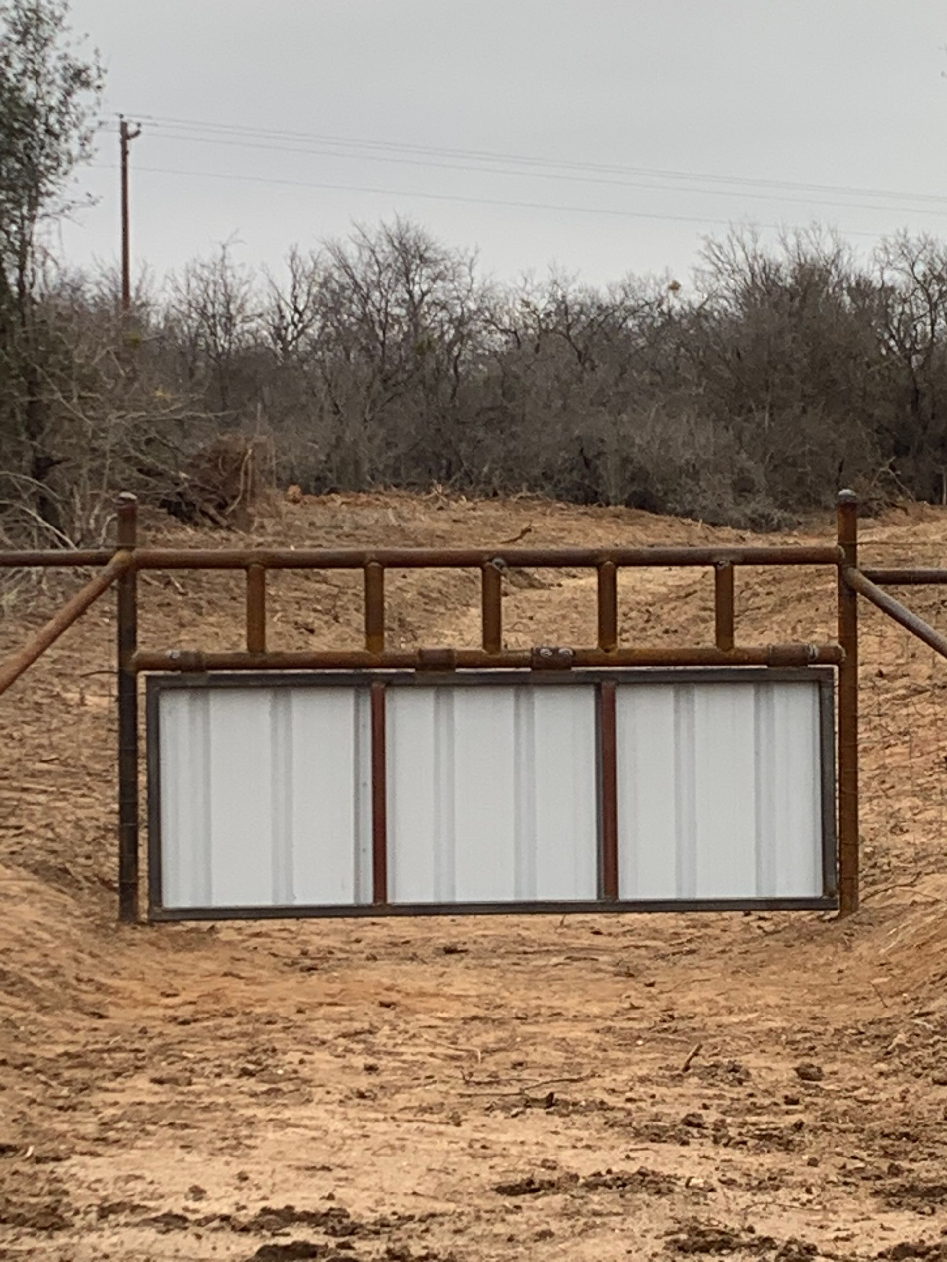 Rusty metal gate with white panels blocking a dirt road in a dry, brushy field