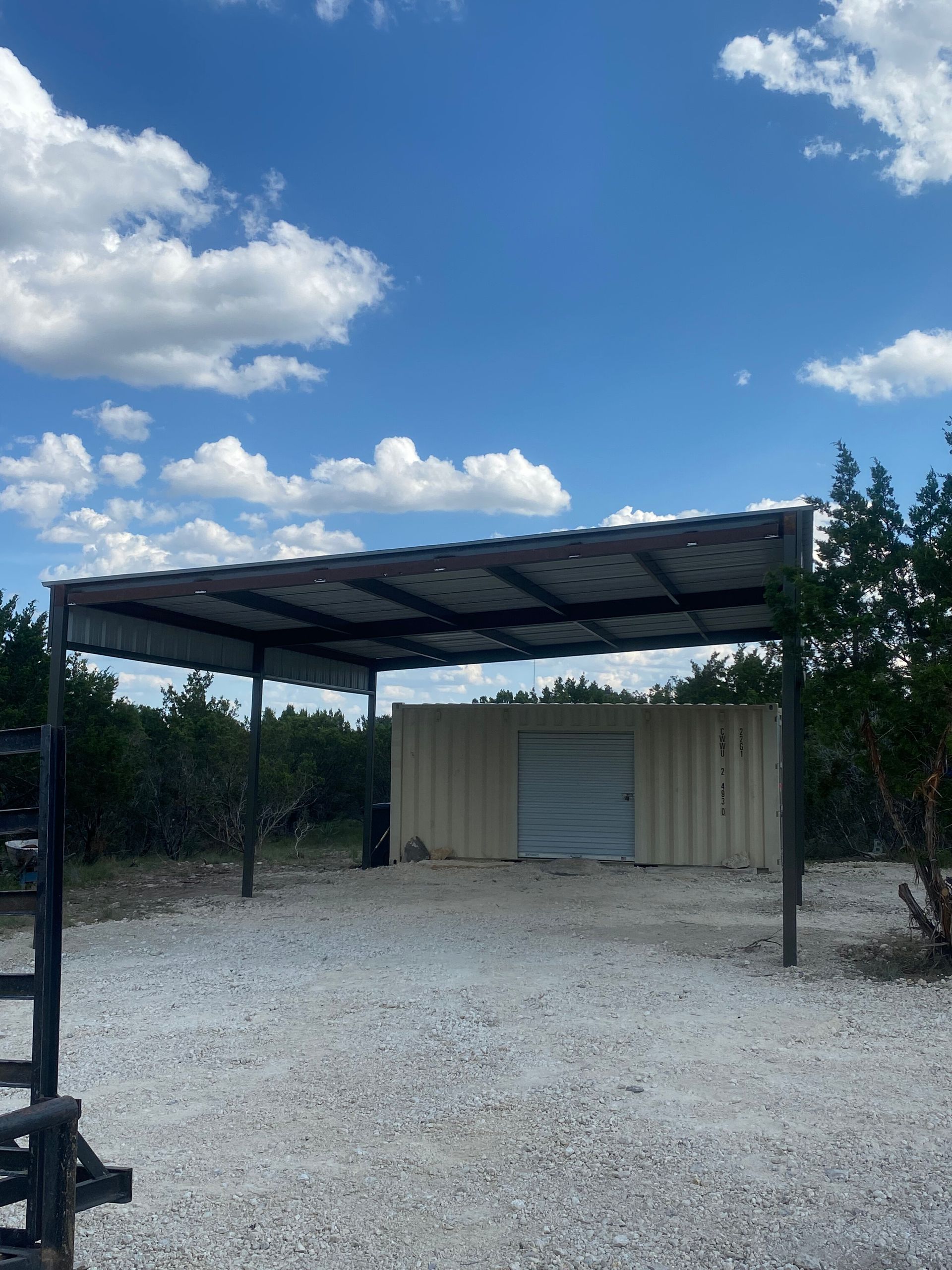 Gravel yard with a metal-roof carport and small beige storage shed under a partly cloudy blue sky