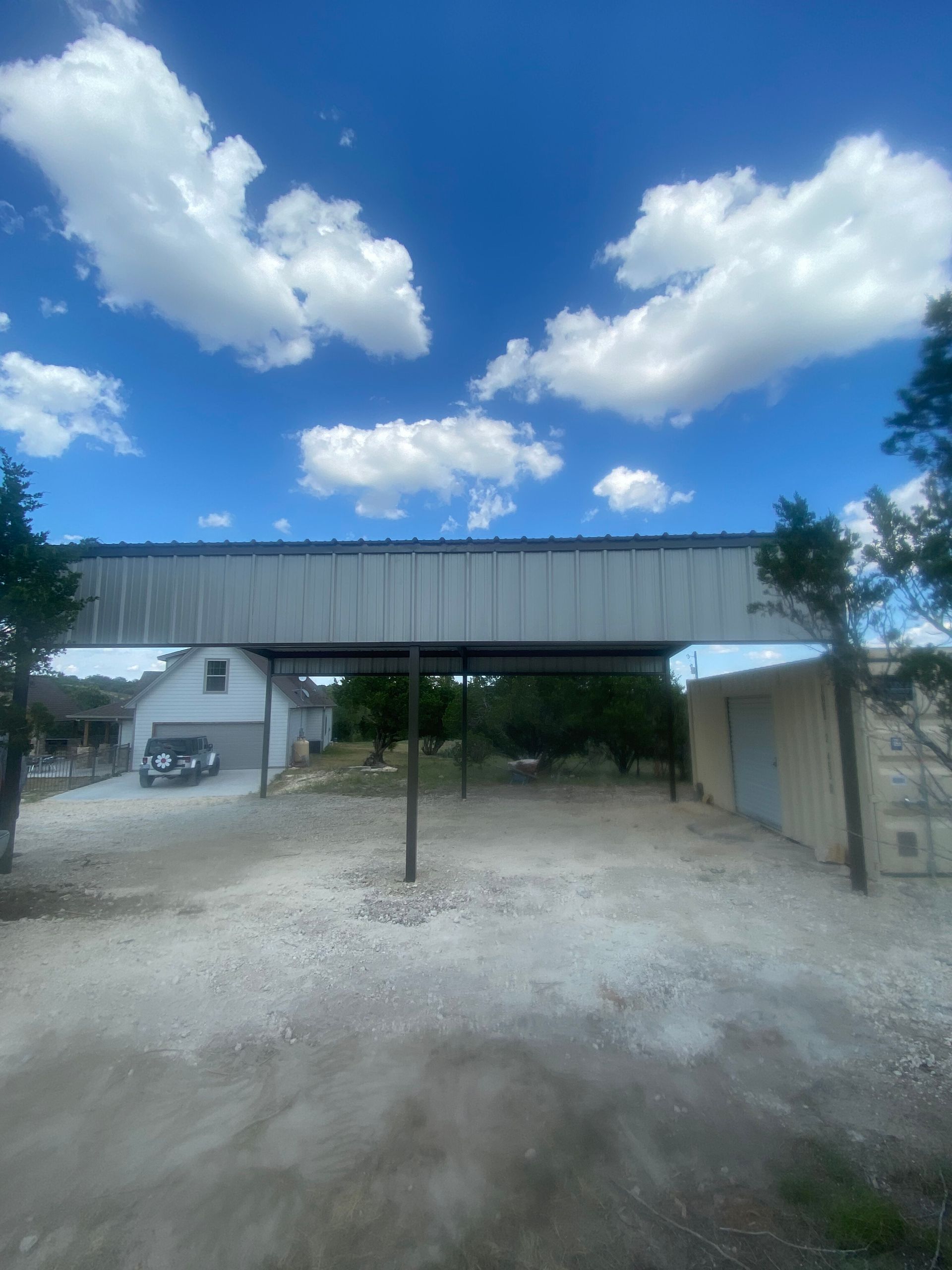 Gravel driveway under a corrugated carport, with a small house and blue sky with clouds in the background