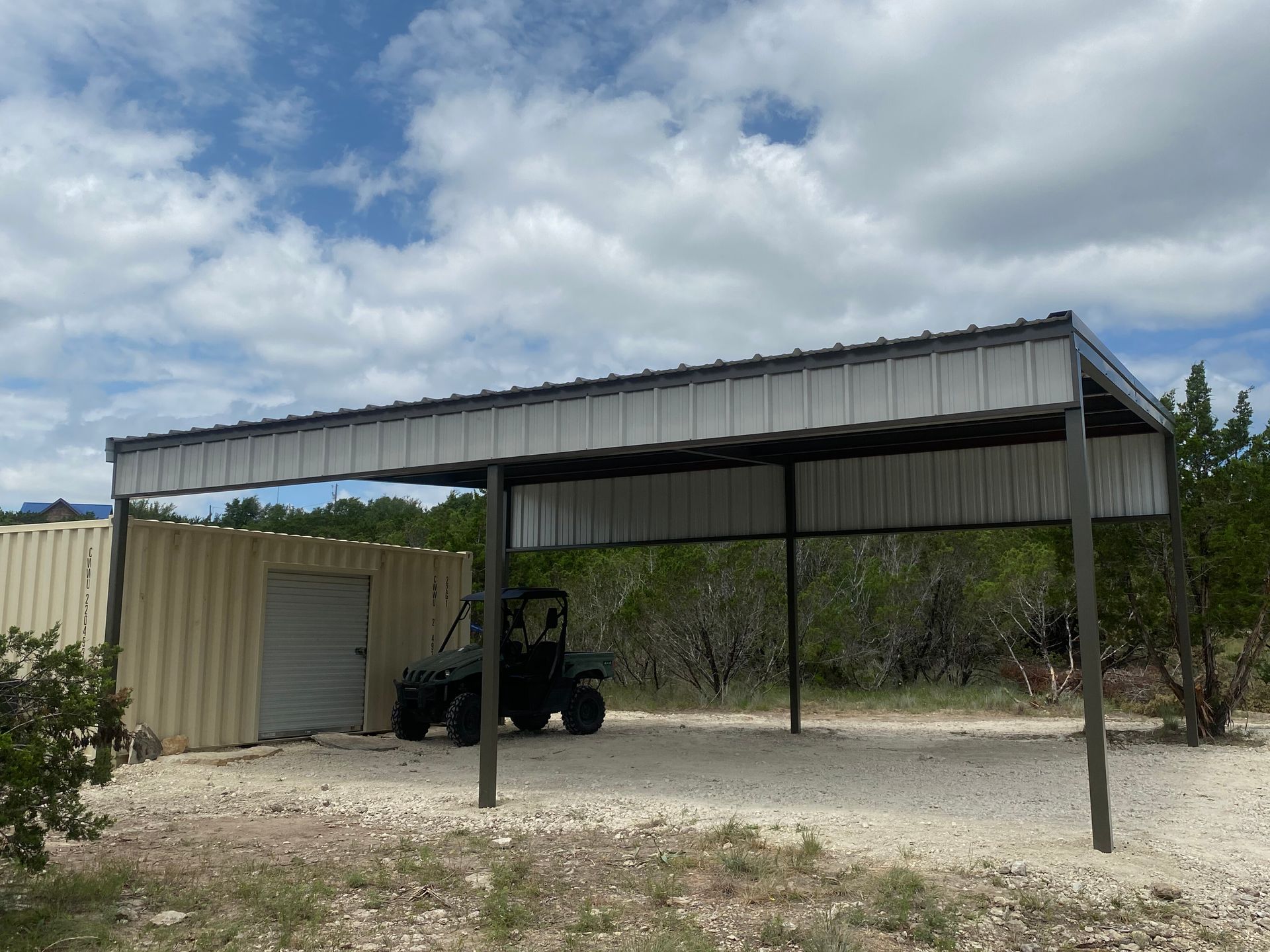 Metal carport with a utility shed and a tractor parked underneath on a gravel lot under a cloudy sky