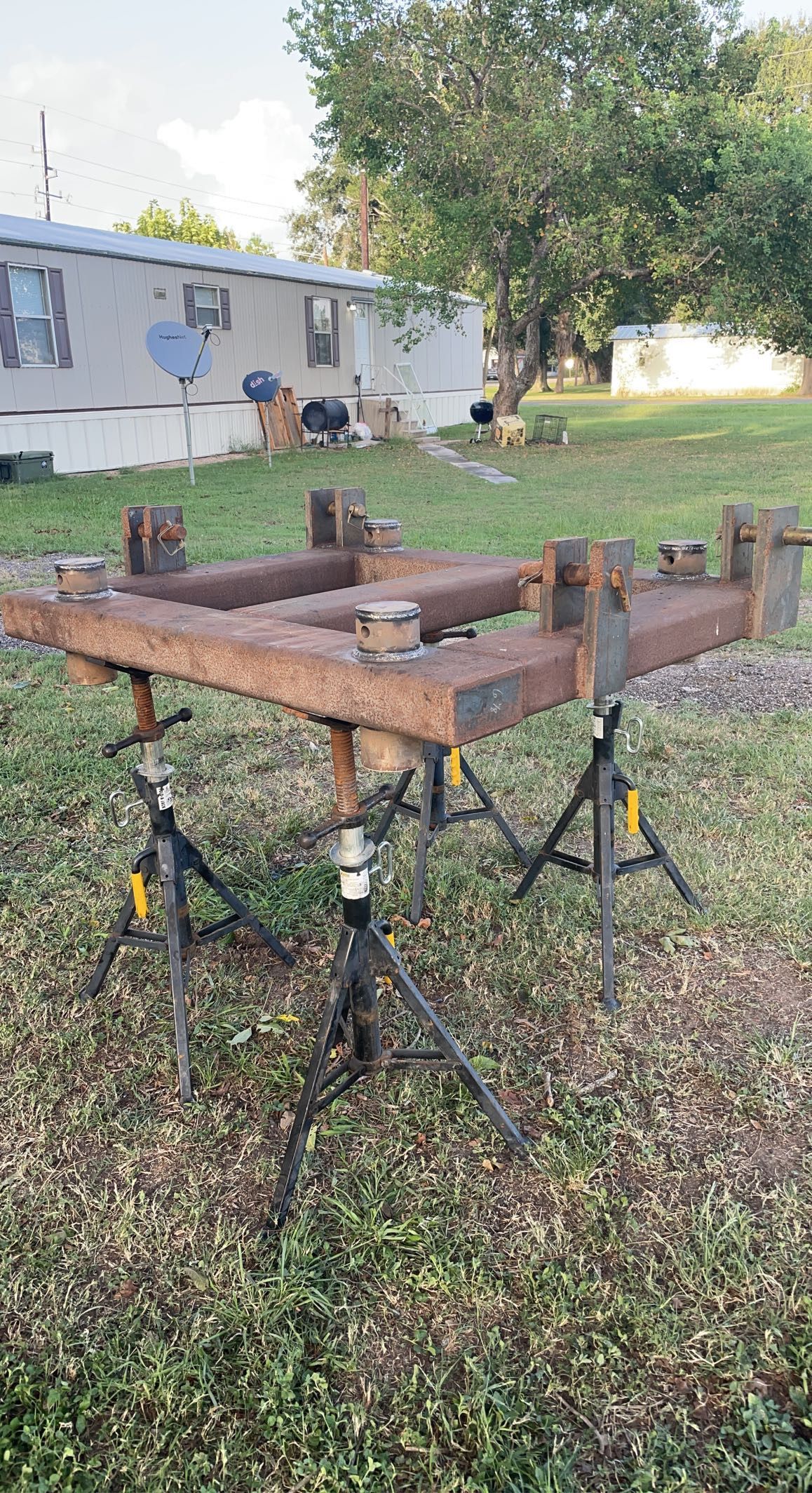 Rusty metal frame on sawhorses in a grassy yard beside a white mobile home and trees