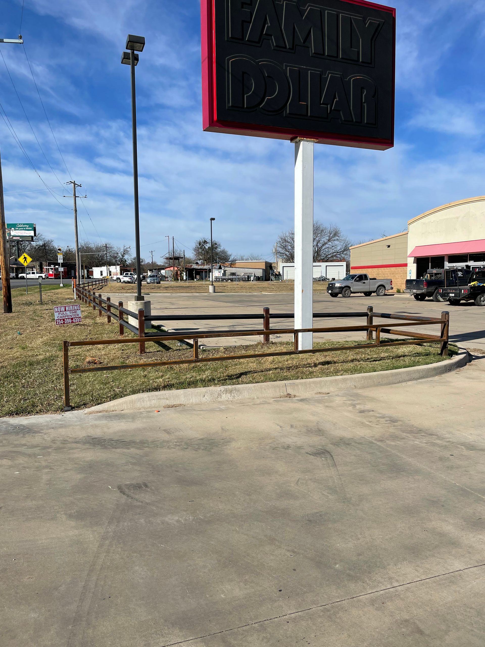 Tall sign by a roadside parking lot under a blue sky, with a wooden fence and storefronts nearby