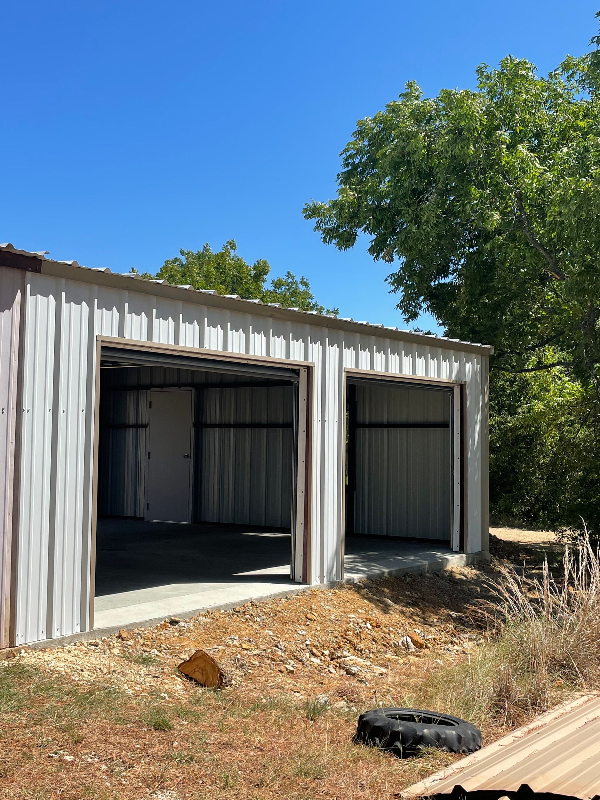 White metal shed with two open bays beside trees, under a clear blue sky.