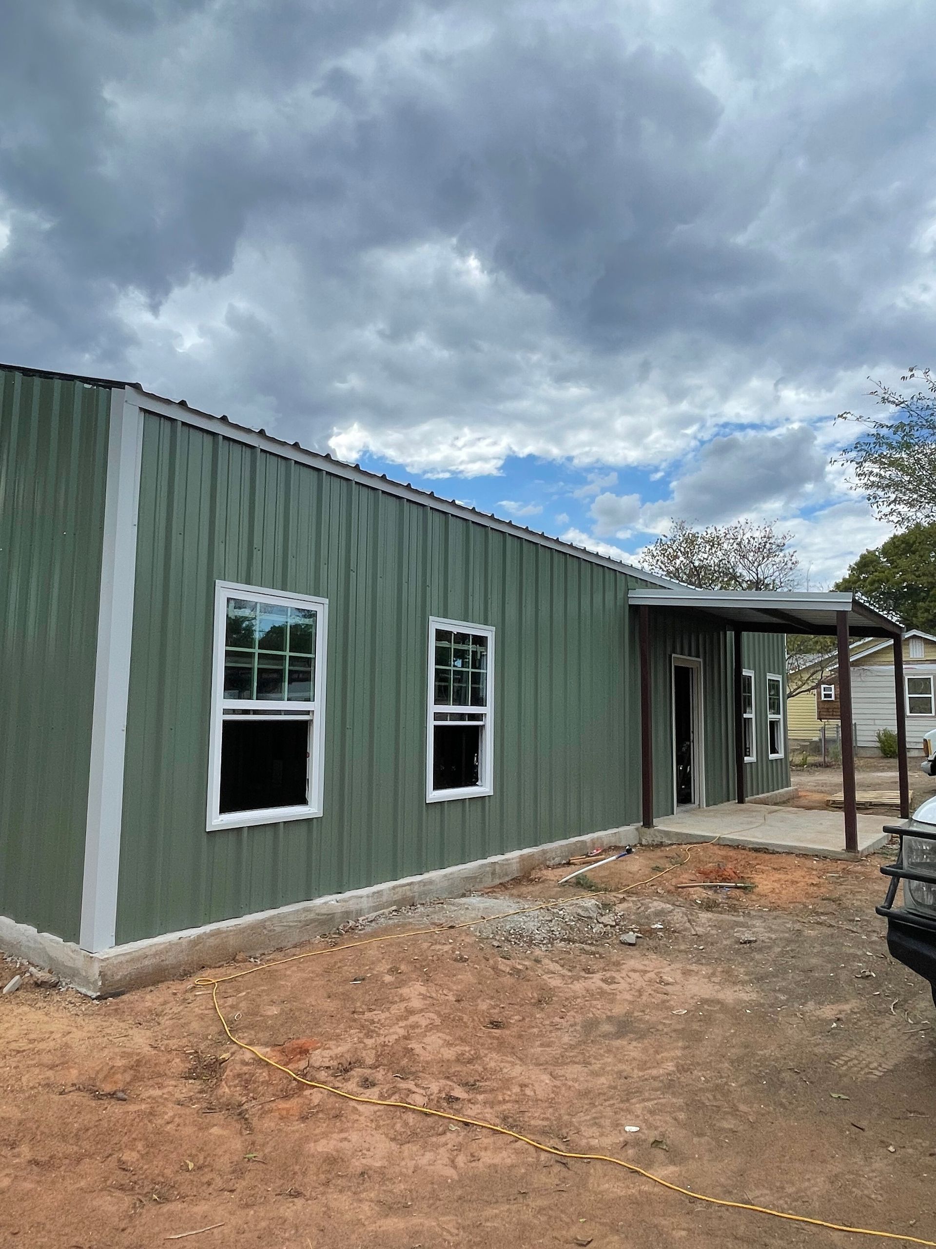Green metal-sided house under construction with two windows and a covered porch on a dirt lot