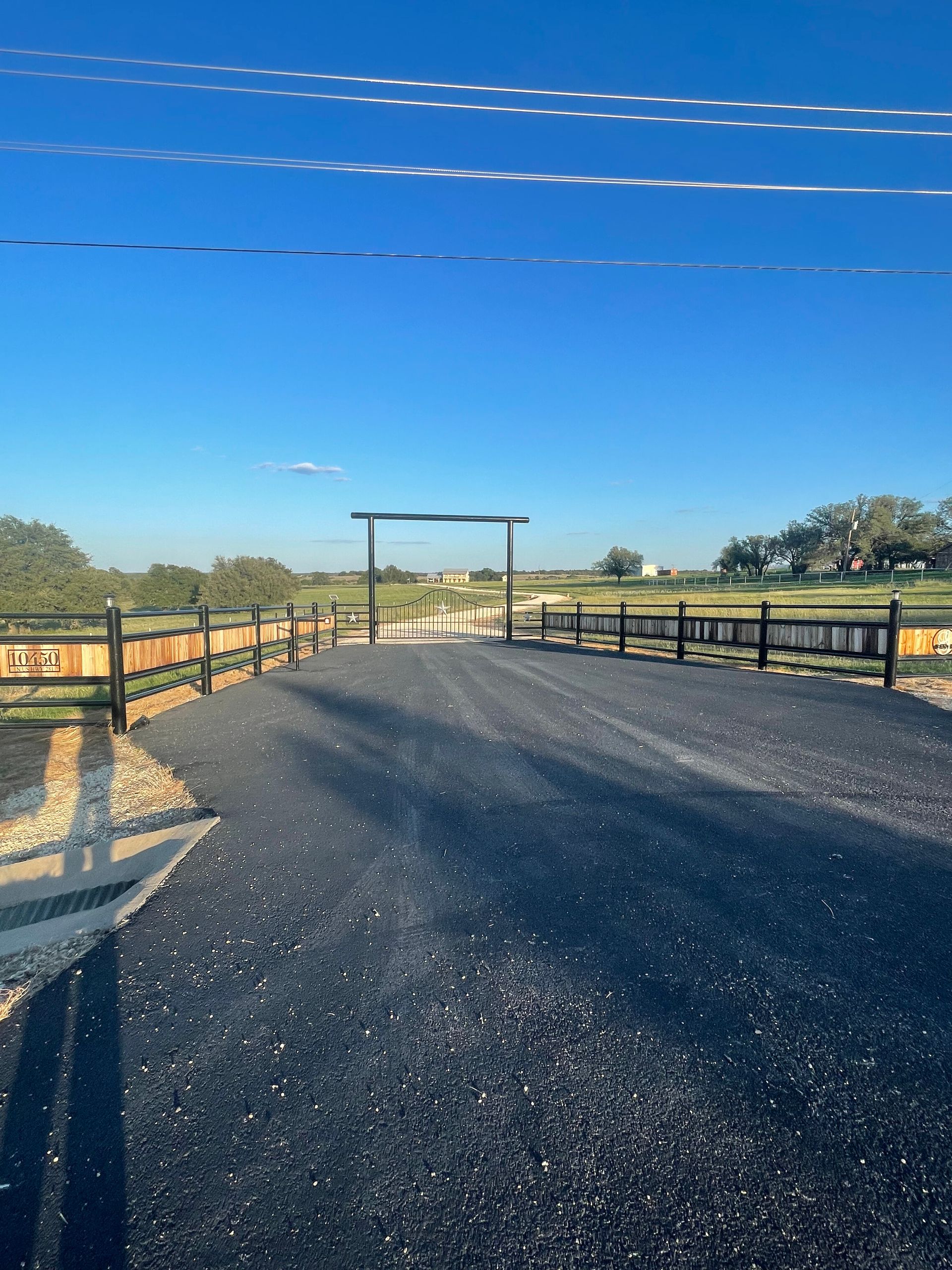 Gravel driveway leading to a wooden gate under a bright blue sky, with fenced fields on both sides.