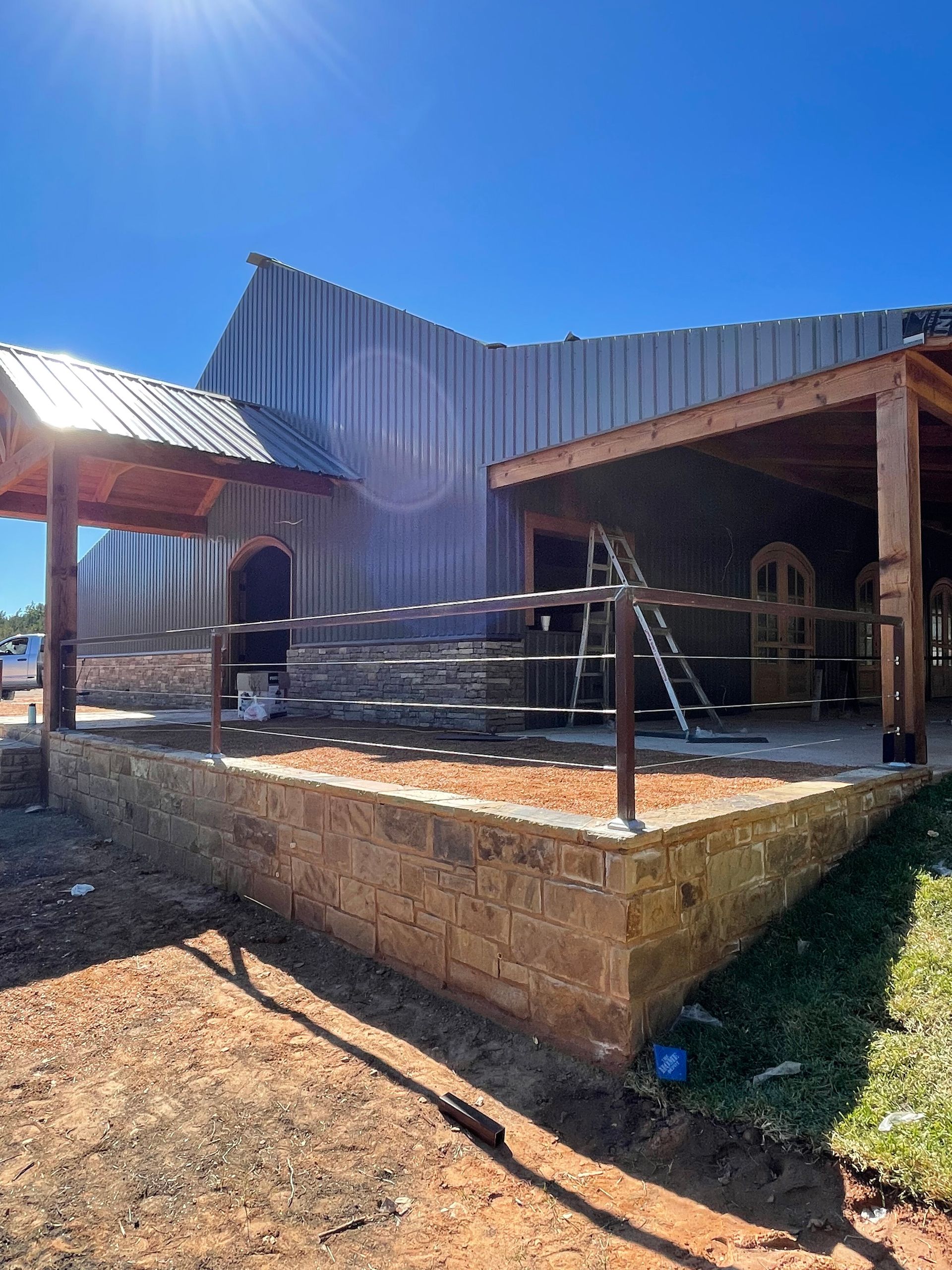 Rustic outdoor patio with wooden beams, corrugated metal roof, and a raised stone platform under a bright blue sky