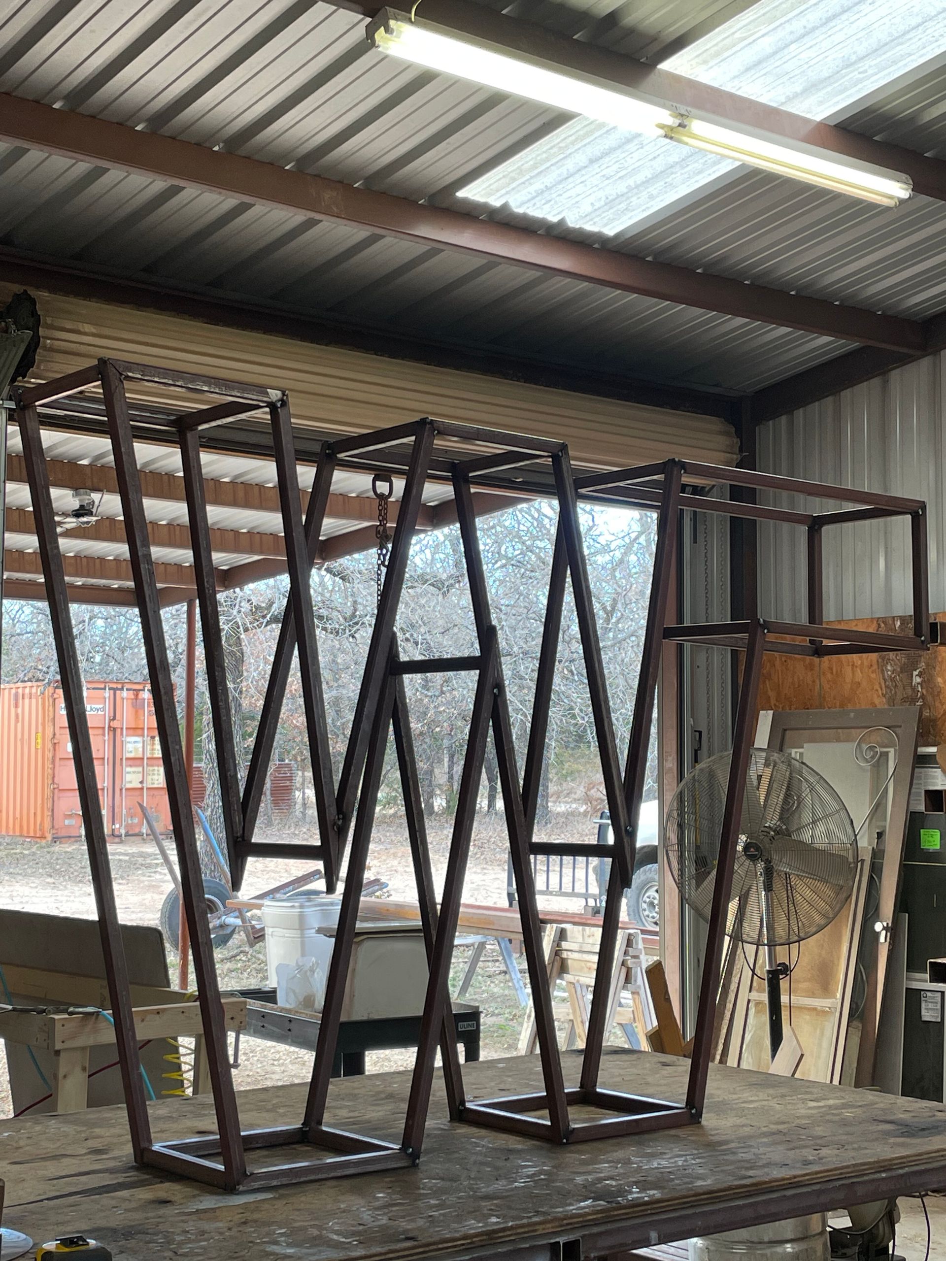 Metal geometric frames arranged on a workbench inside a workshop with a corrugated roof.