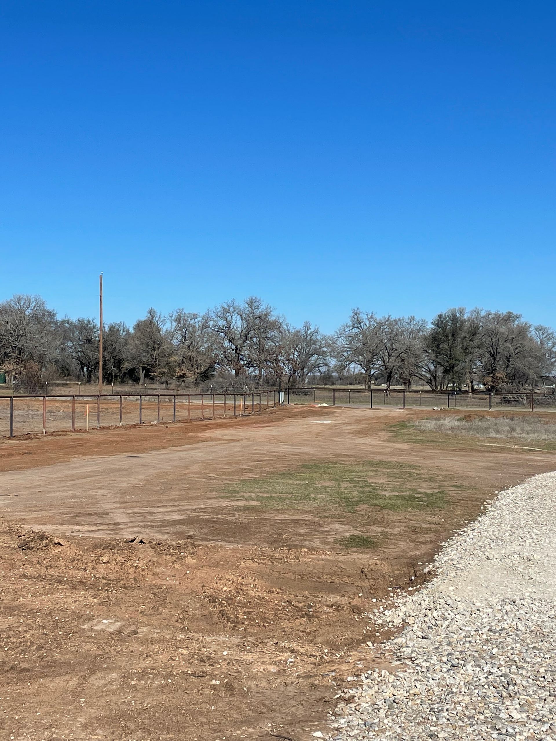 Open dirt field with wooden fence, leafless trees, and bright blue sky