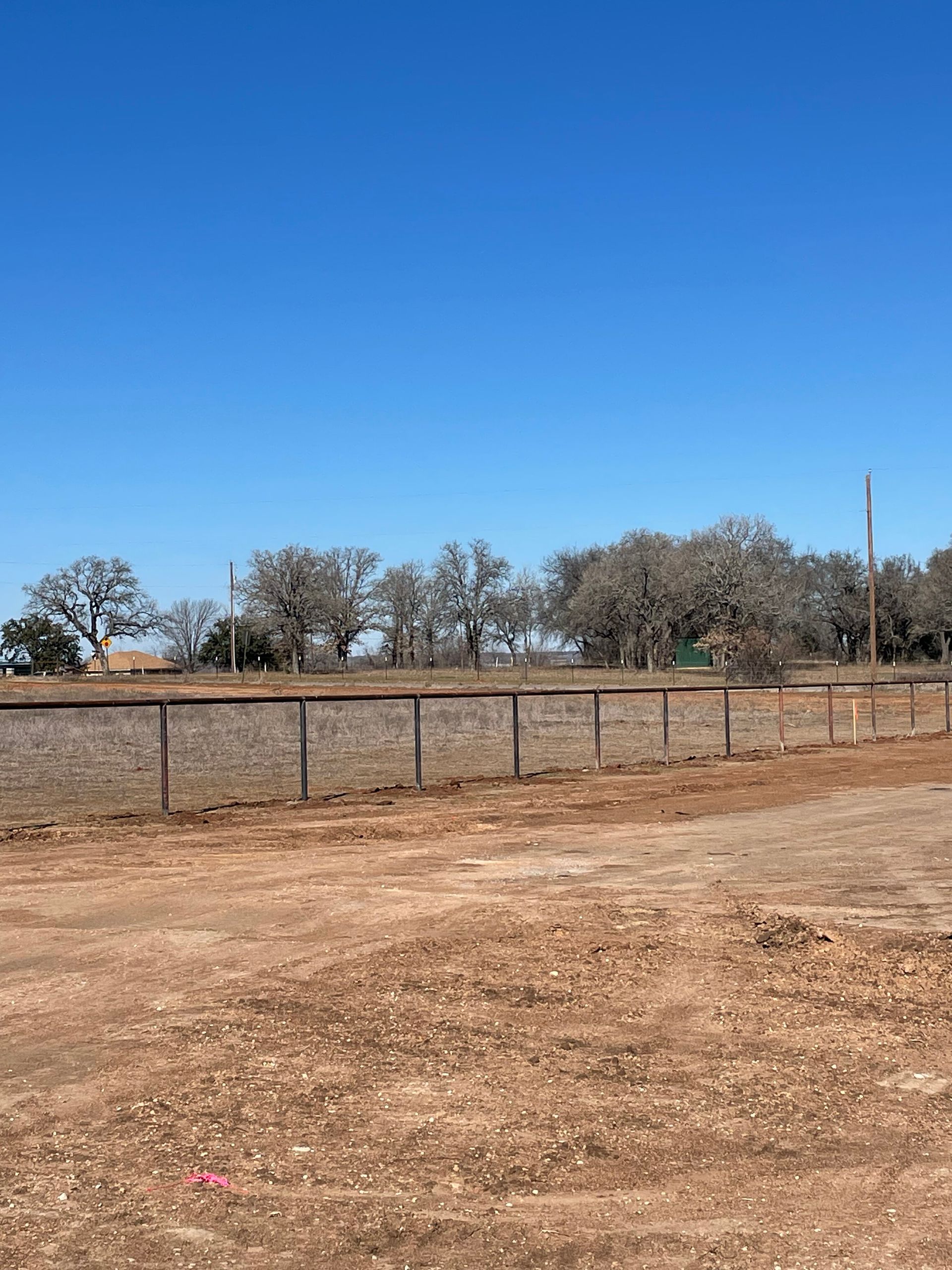 Dry dirt field with a wire fence, sparse trees, and a clear blue sky.