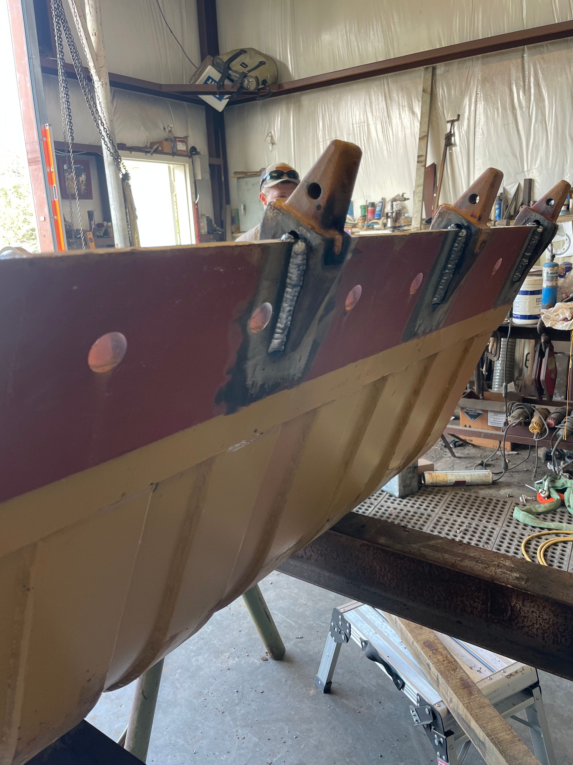 Brown metal boat hull in a workshop, with clamps and tools attached during repair.
