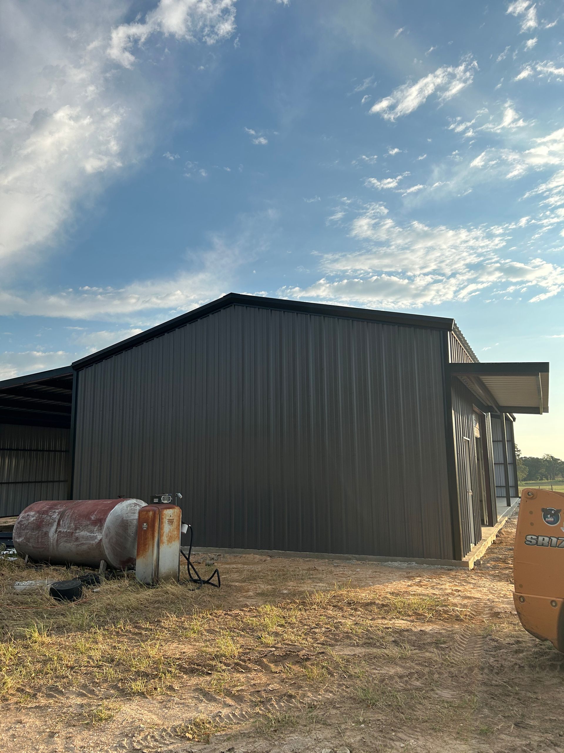 Dark metal farm building with a cloudy sky, dry ground, and farm equipment in the foreground