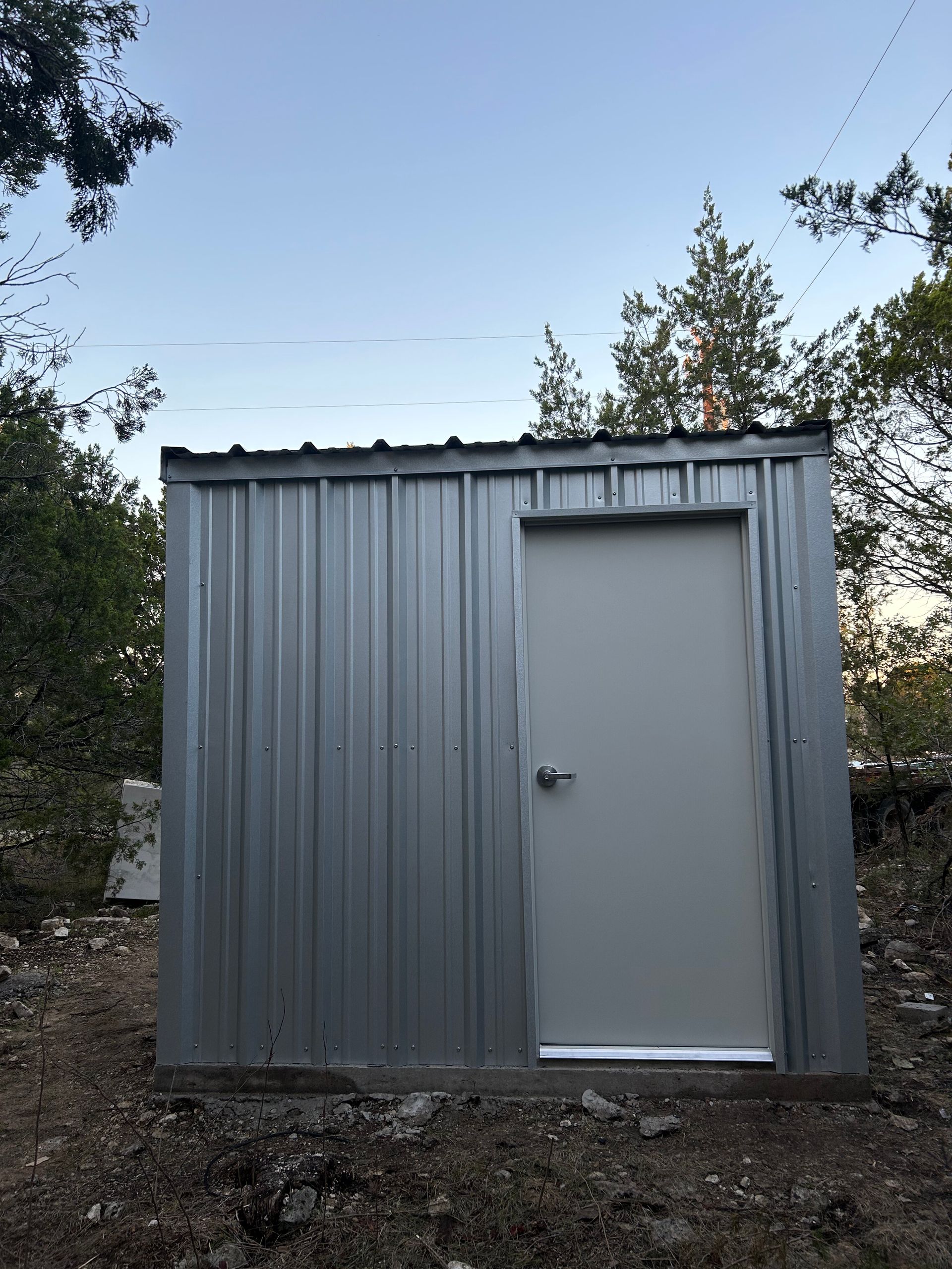 Small gray metal shed with a single door in a wooded outdoor area.