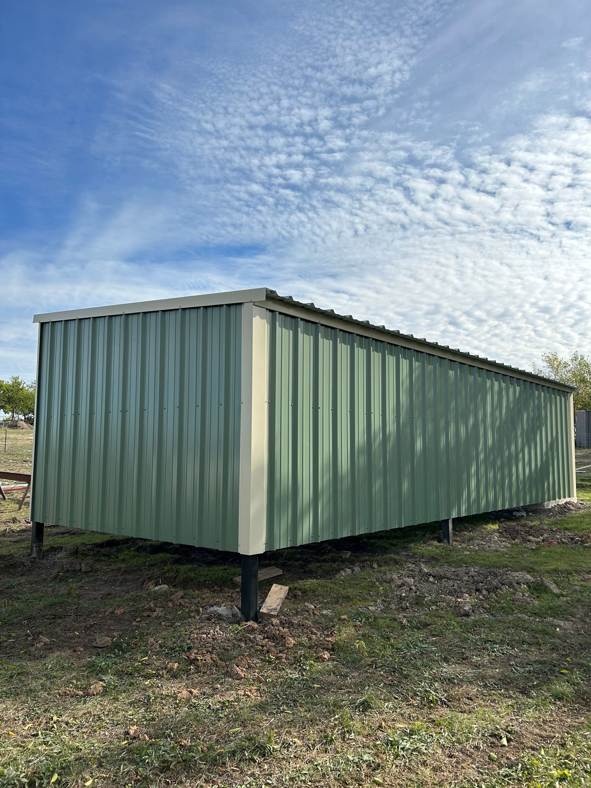 Green corrugated metal shed on short stilts in a grassy field under a cloudy blue sky