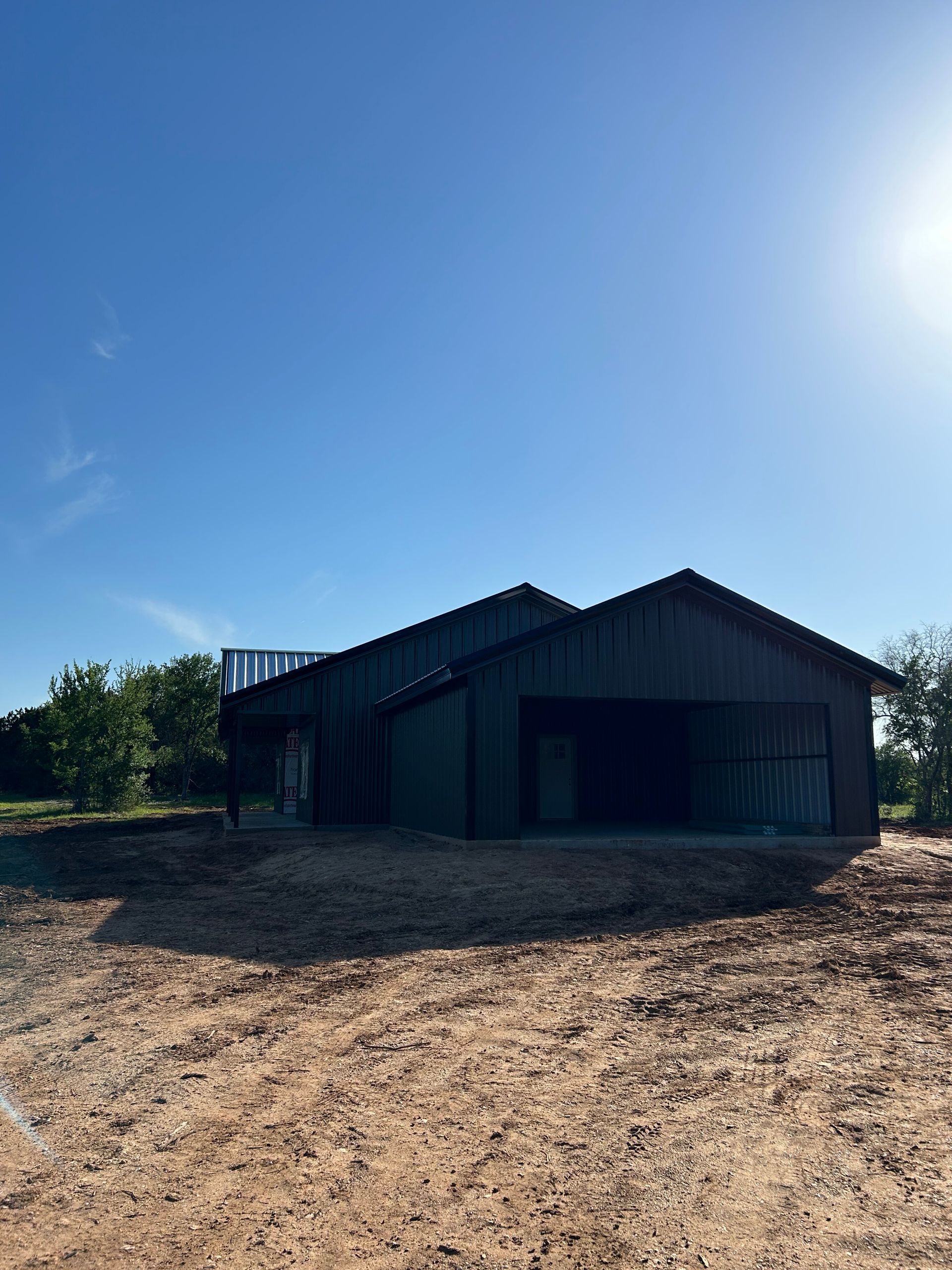 Dark metal barn on a gravel lot under a bright blue sky with the sun at upper right