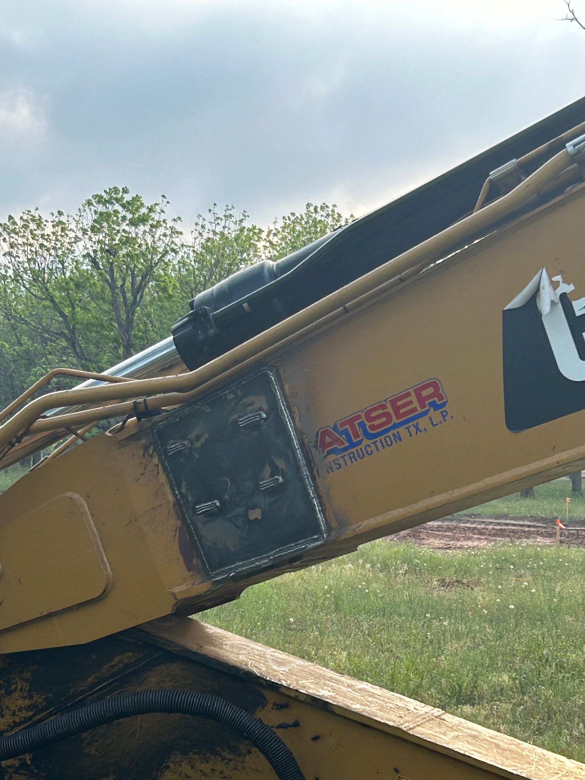 Close-up of a yellow excavator boom with hydraulic hoses and a logo against a grassy outdoor background