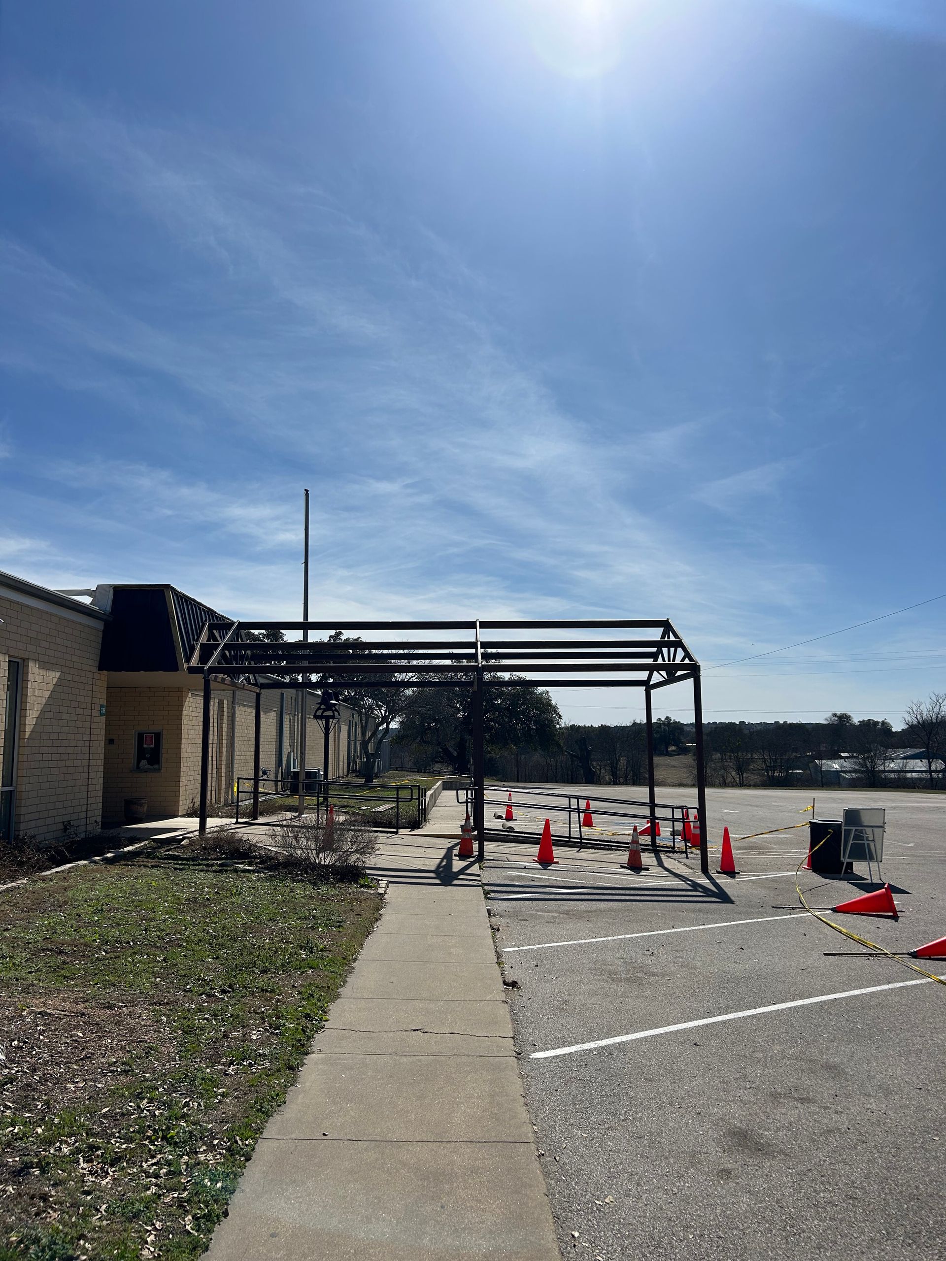 Outdoor construction area with canopy frame, traffic cones, and a concrete path beside a building.