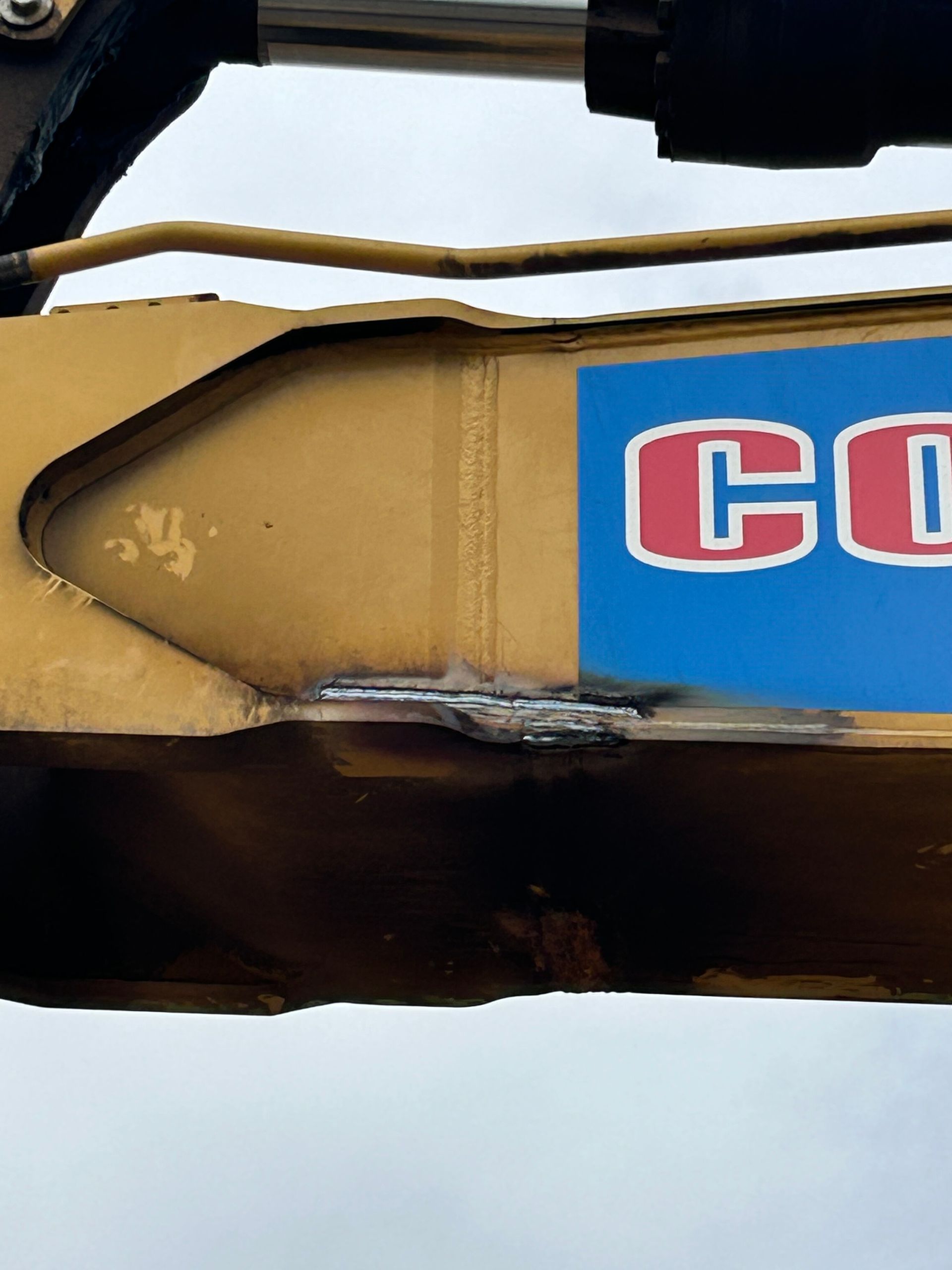 Close-up of a yellow construction machine arm with a blue logo panel and black hydraulic hose.