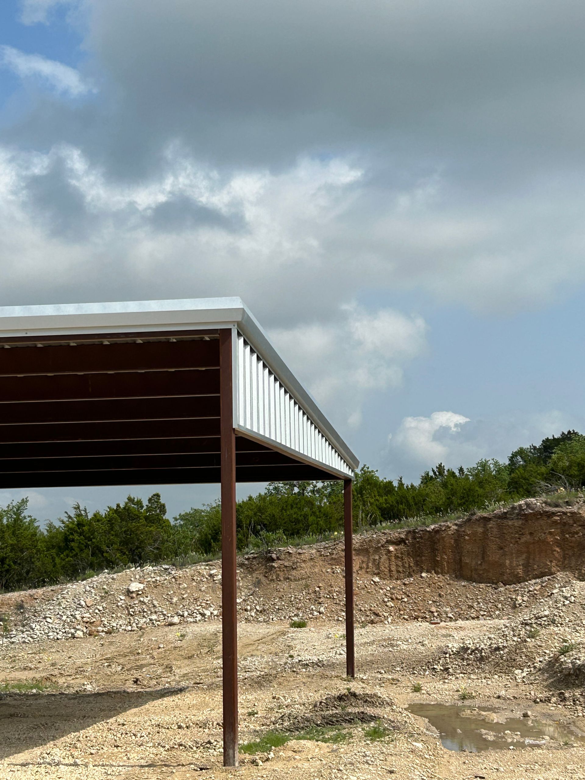 Rusty metal canopy on posts in a rocky outdoor lot under a cloudy sky