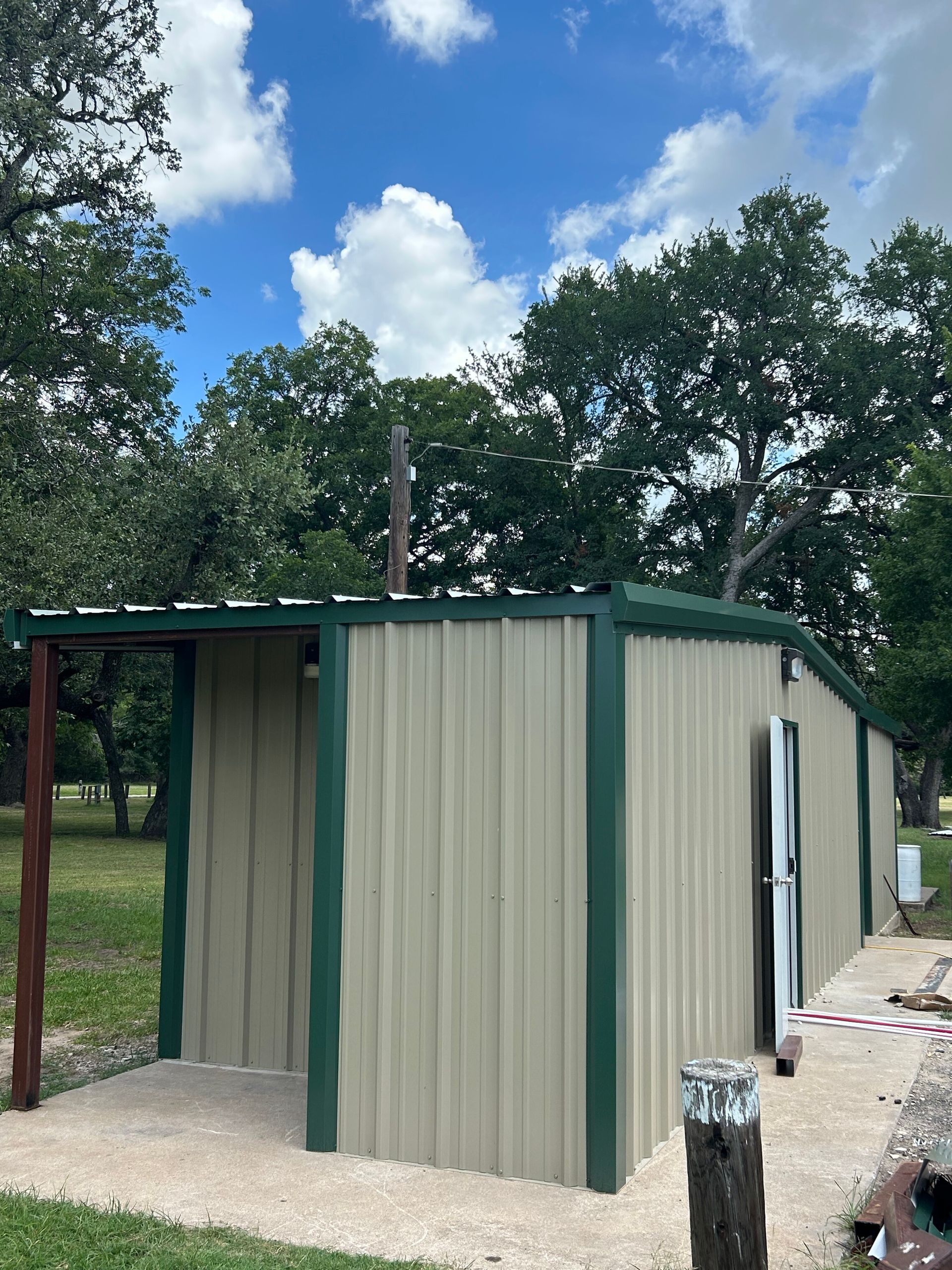 Green-and-beige small building or shed with a metal roof on a gravel pad under trees and blue sky.