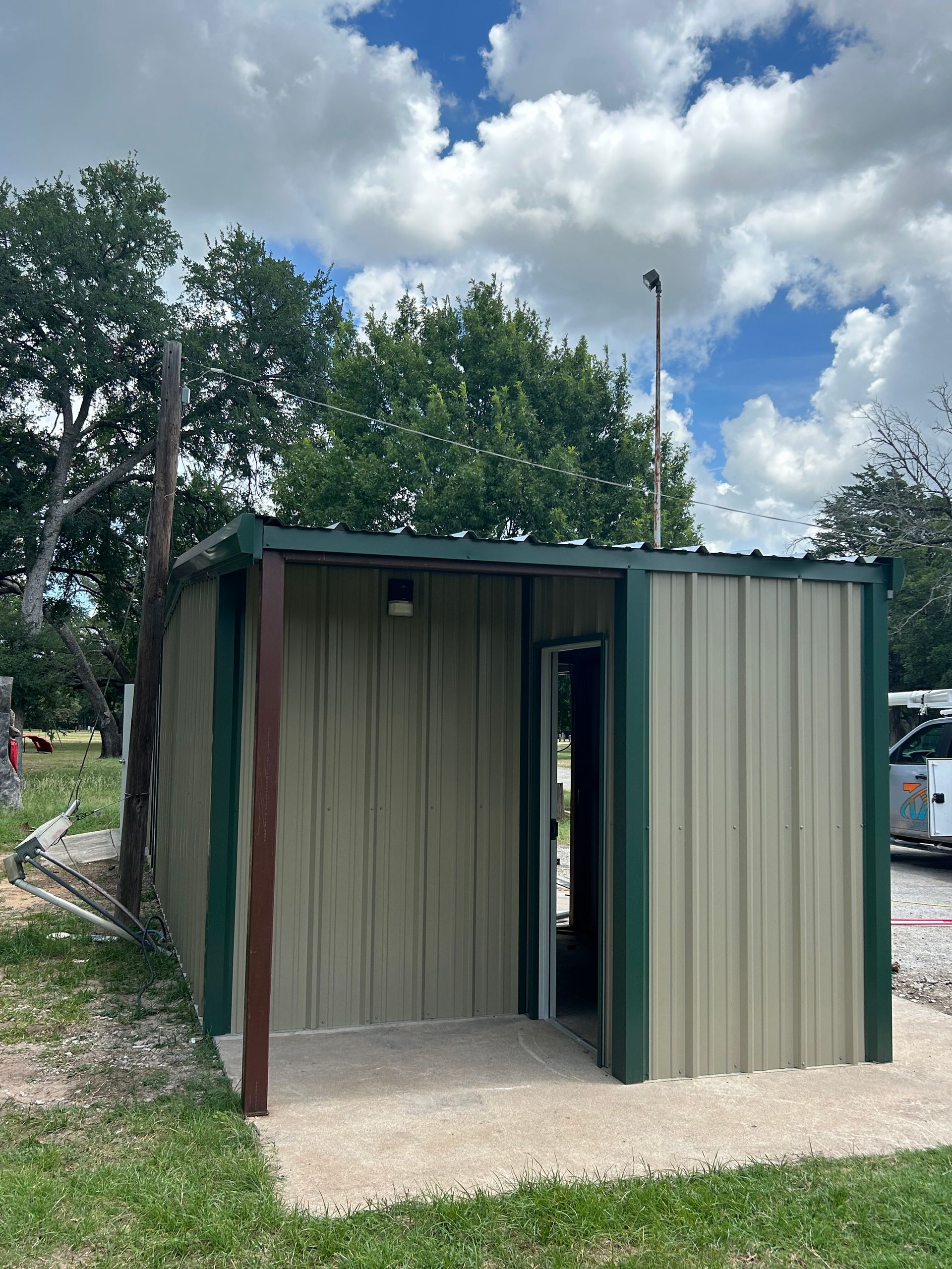 Small green metal shed with an open doorway on a concrete pad in a grassy yard under trees and cloudy sky